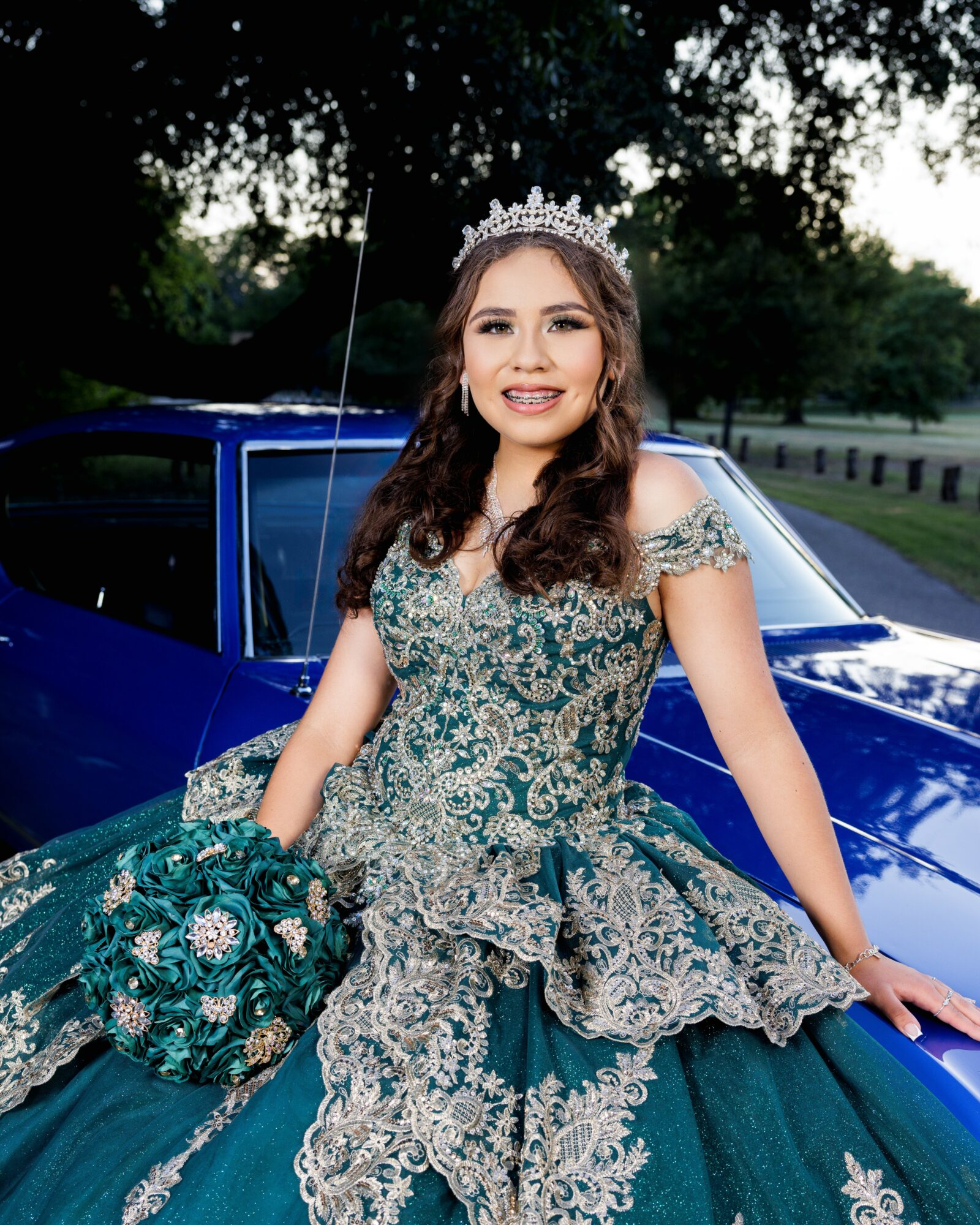 Young woman in an ornate teal and silver gown with a tiara, standing outdoors next to a blue car, smiling.