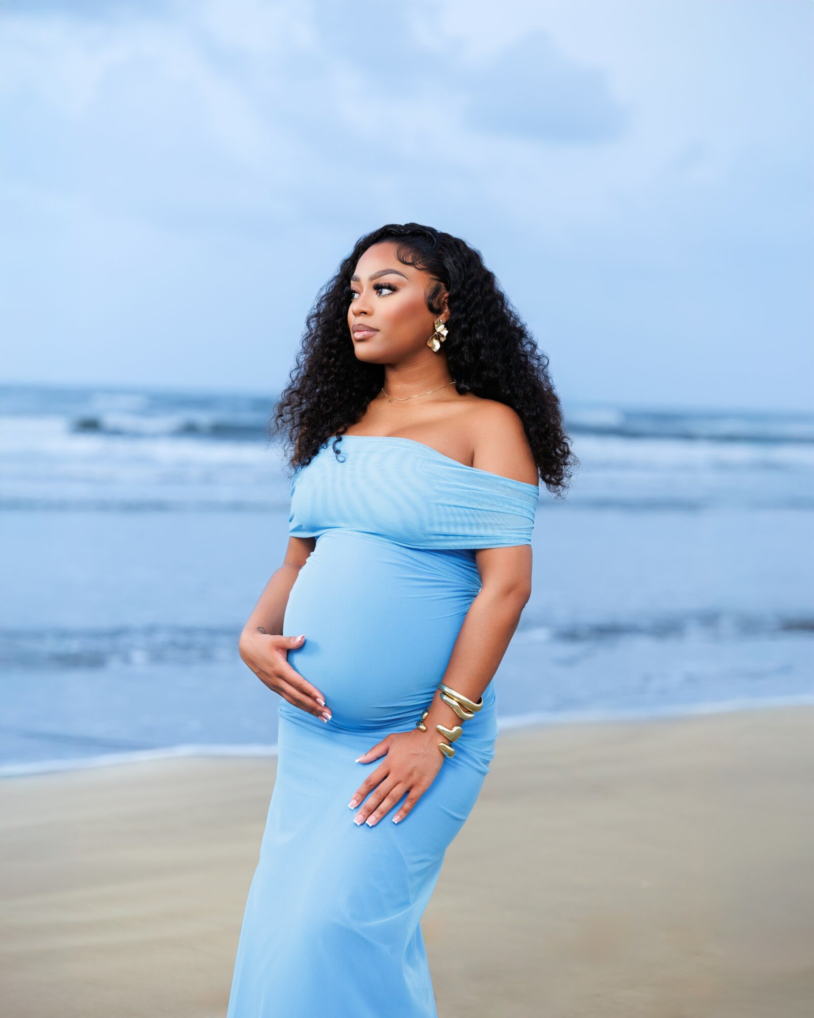 Pregnant woman in a blue dress standing on the beach, holding her belly, with ocean and sky in background.