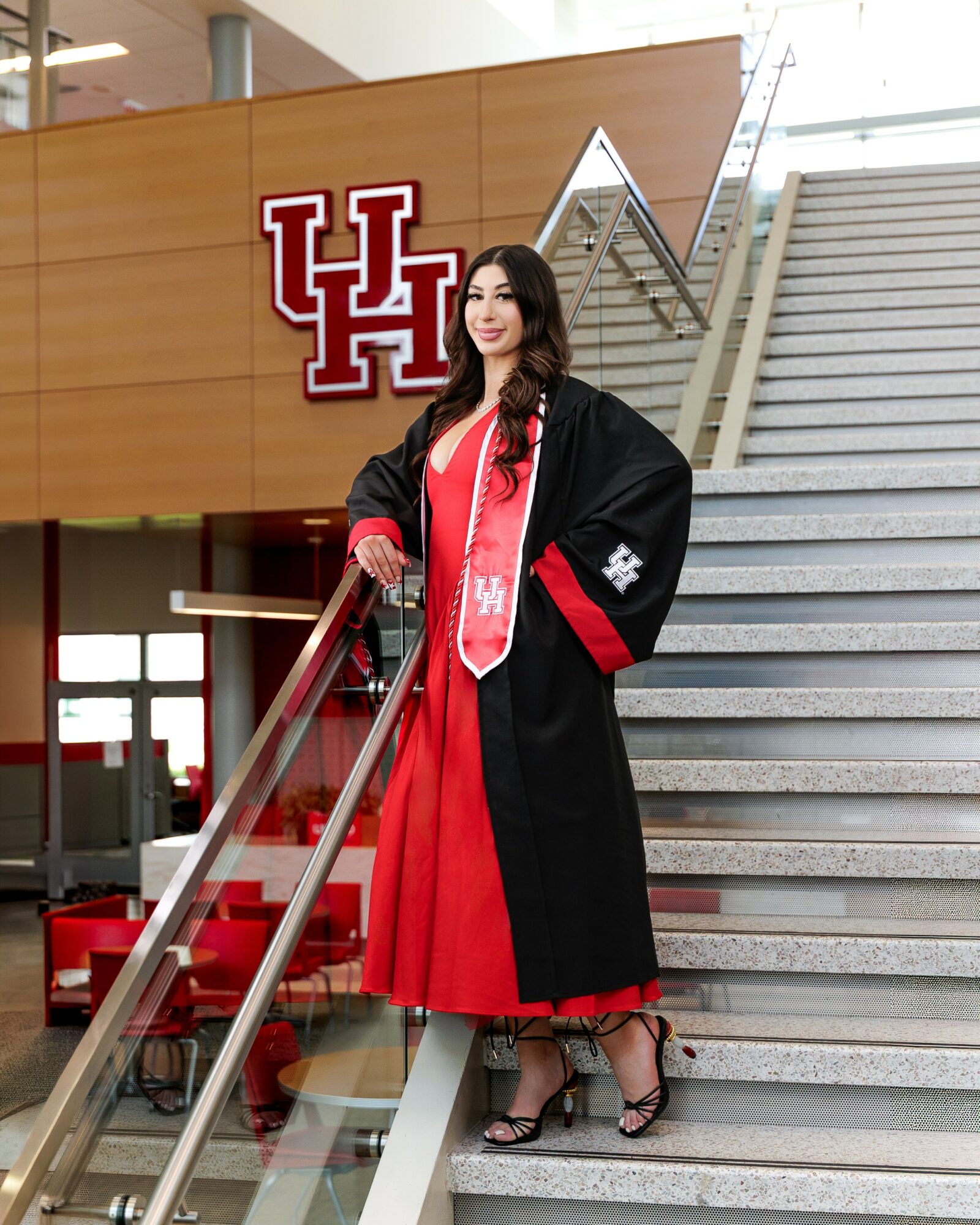 Woman in graduation gown and red dress standing on stairs inside building with University of Houston logo behind her.