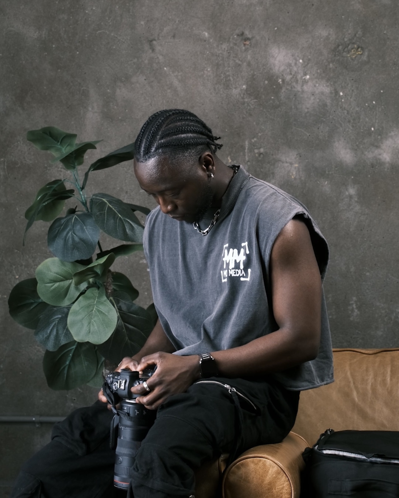 Person with braided hair sitting on a couch, looking at a camera, next to a plant and a bag.