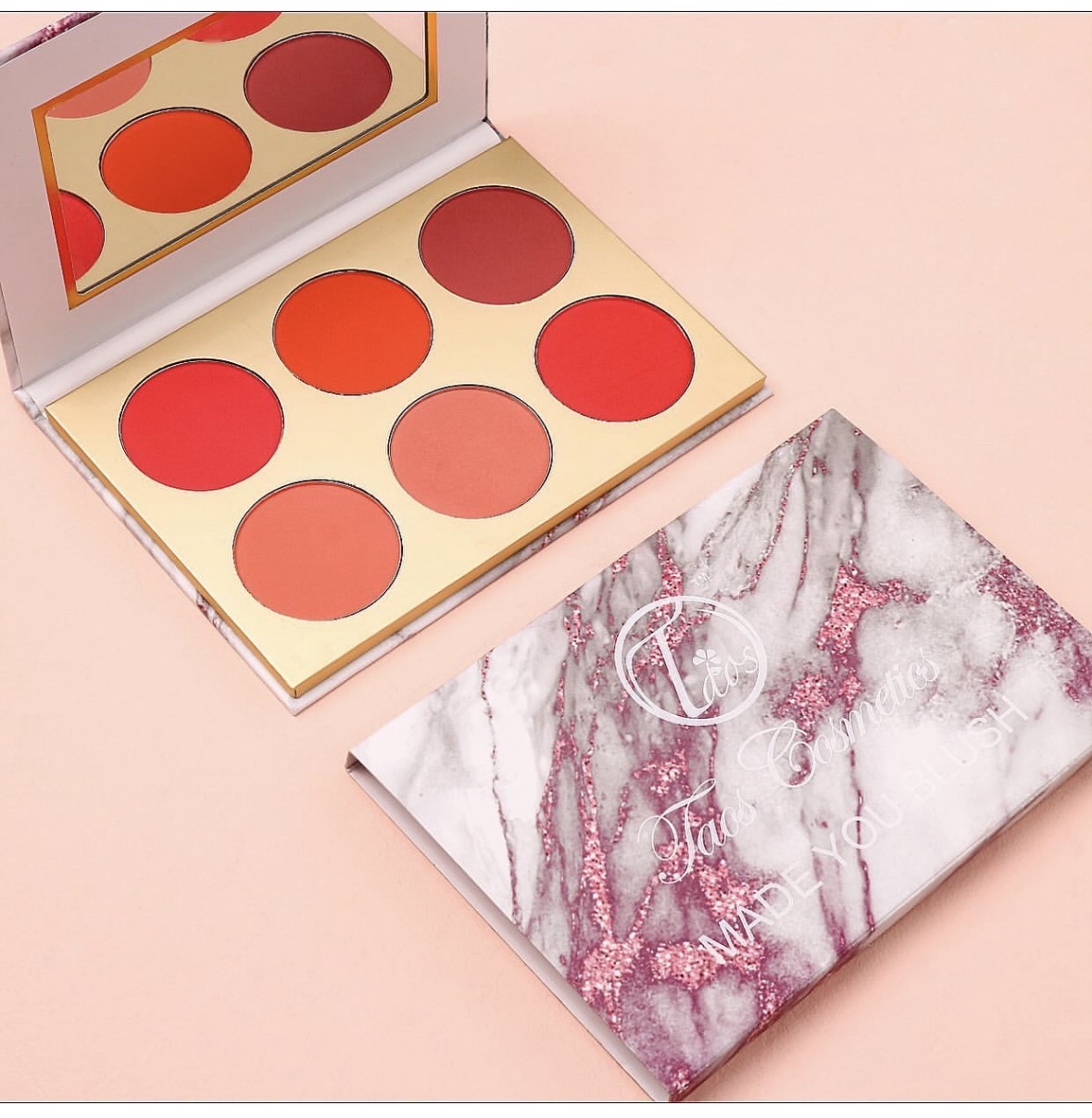 Open makeup palette with six red and orange shades, marble-patterned case nearby, on pink background.