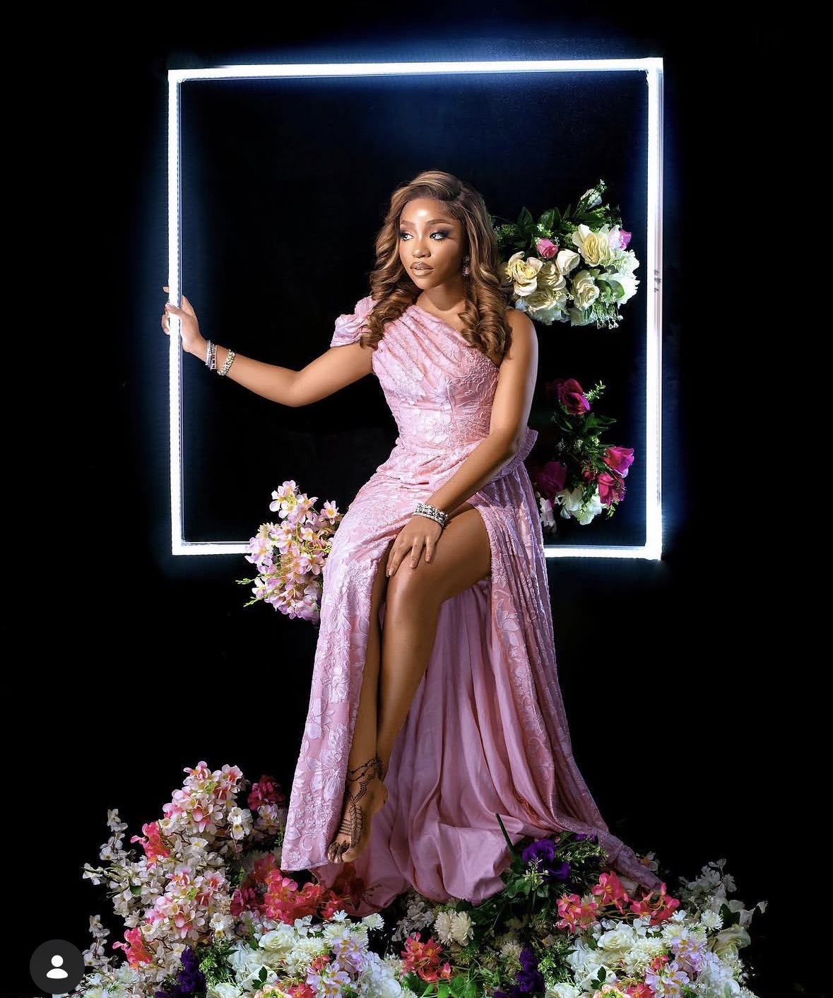 Young woman in pink gown sitting on a floral arrangement, framed by a glowing square, with flowers around her and behind her.