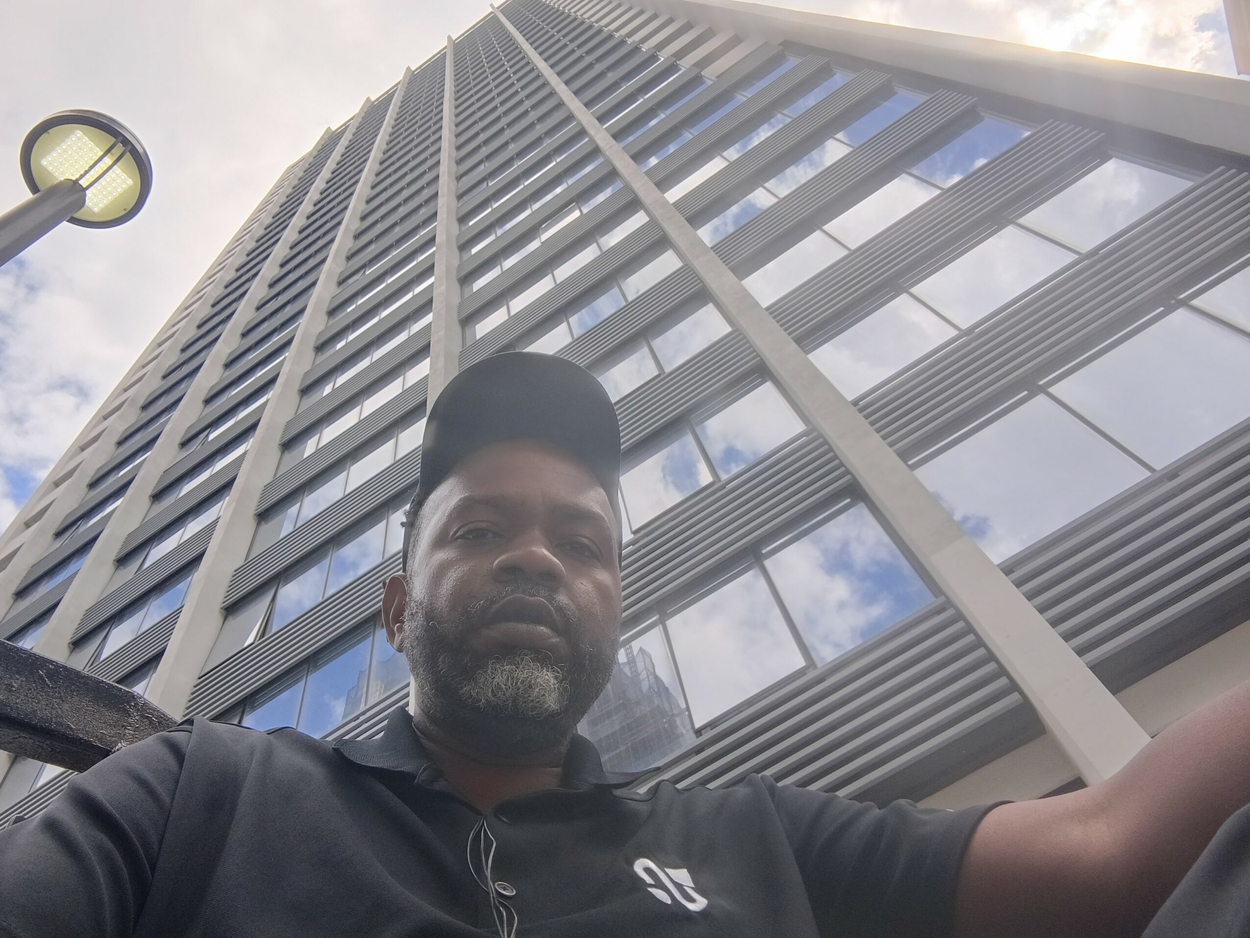 A man with a beard and cap taking a selfie in front of a tall modern building with glass windows.