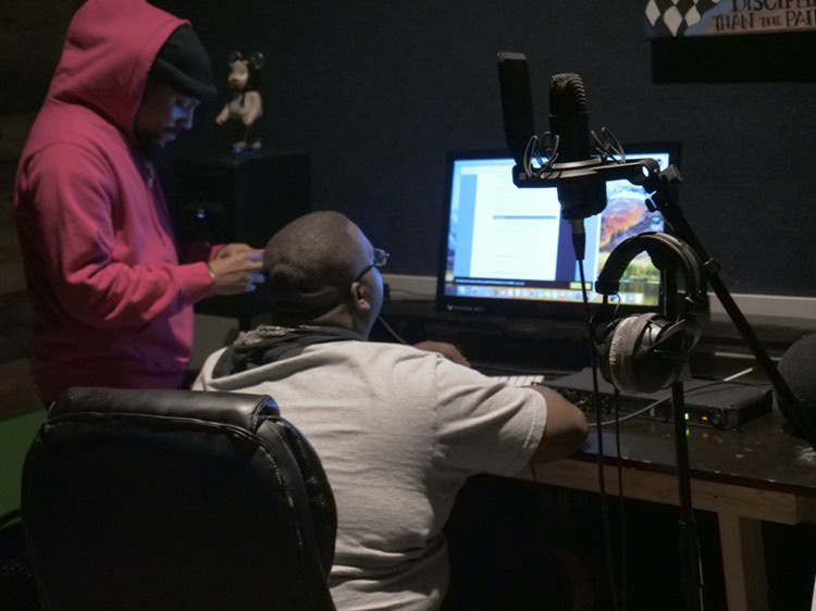 Two people working at a desk with a computer and microphone, one seated and one standing, in a dark room.