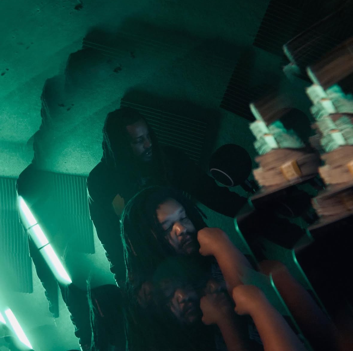 Two people with dreadlocks in a dimly lit room with green lighting, one smiling and the other looking down, with bottles and stacks of money nearby.