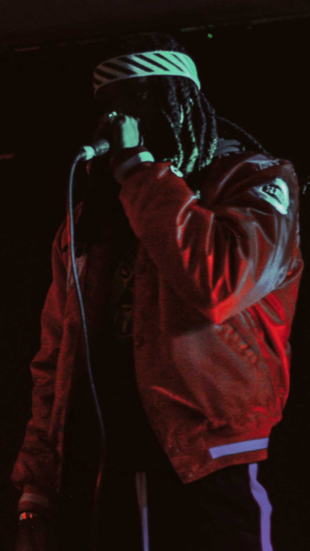 Person with dreadlocks wearing a striped hat and red jacket, holding a microphone in a dark setting.