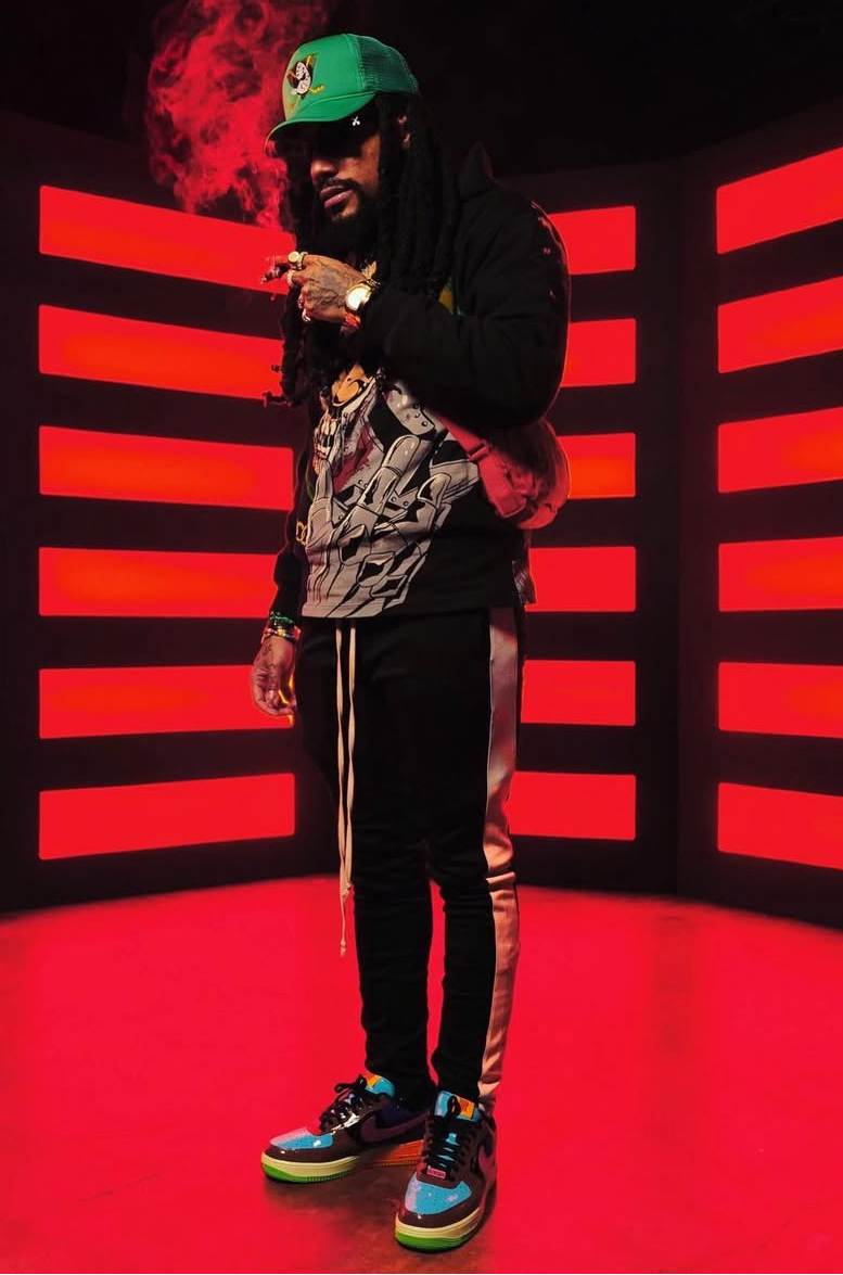 Person standing in front of red-lit background, wearing a green helmet, black jacket, striped pants, and colorful sneakers, smoking.