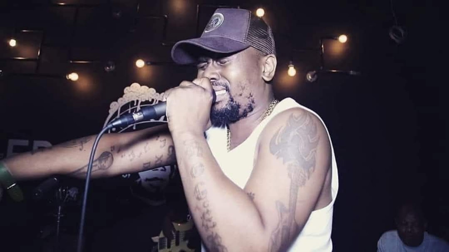 Man singing into a microphone with a cap, tattoos visible on arms, in a dimly lit setting.
