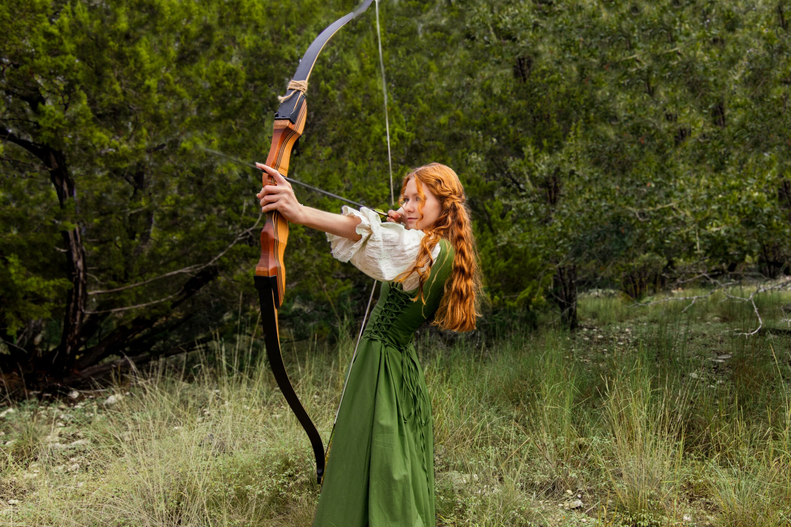 A woman with long red hair in a green dress aims a bow and arrow outdoors in a grassy area with trees.