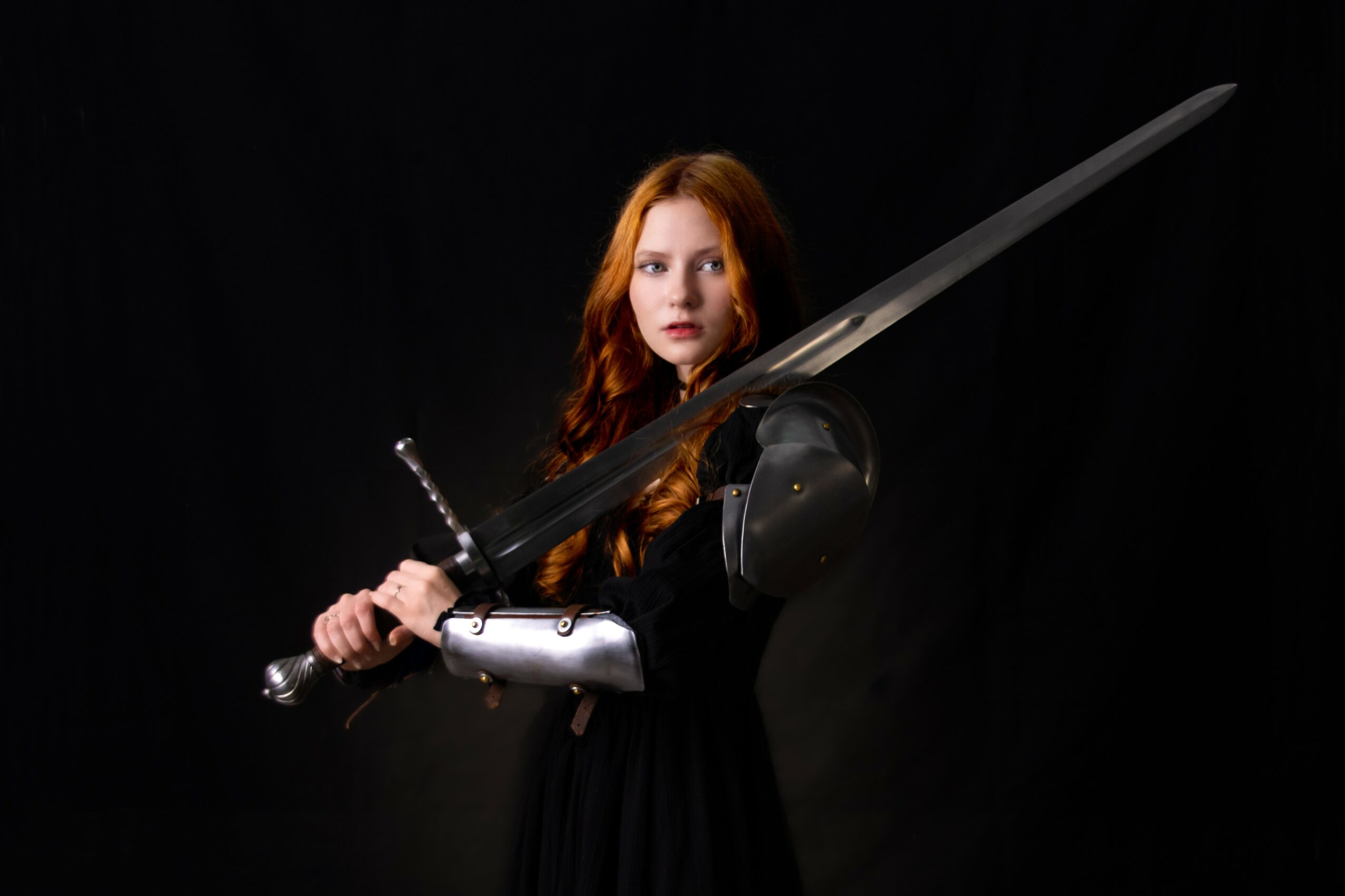 A woman with long red hair holds a sword and shield, dressed in black, against a dark background.