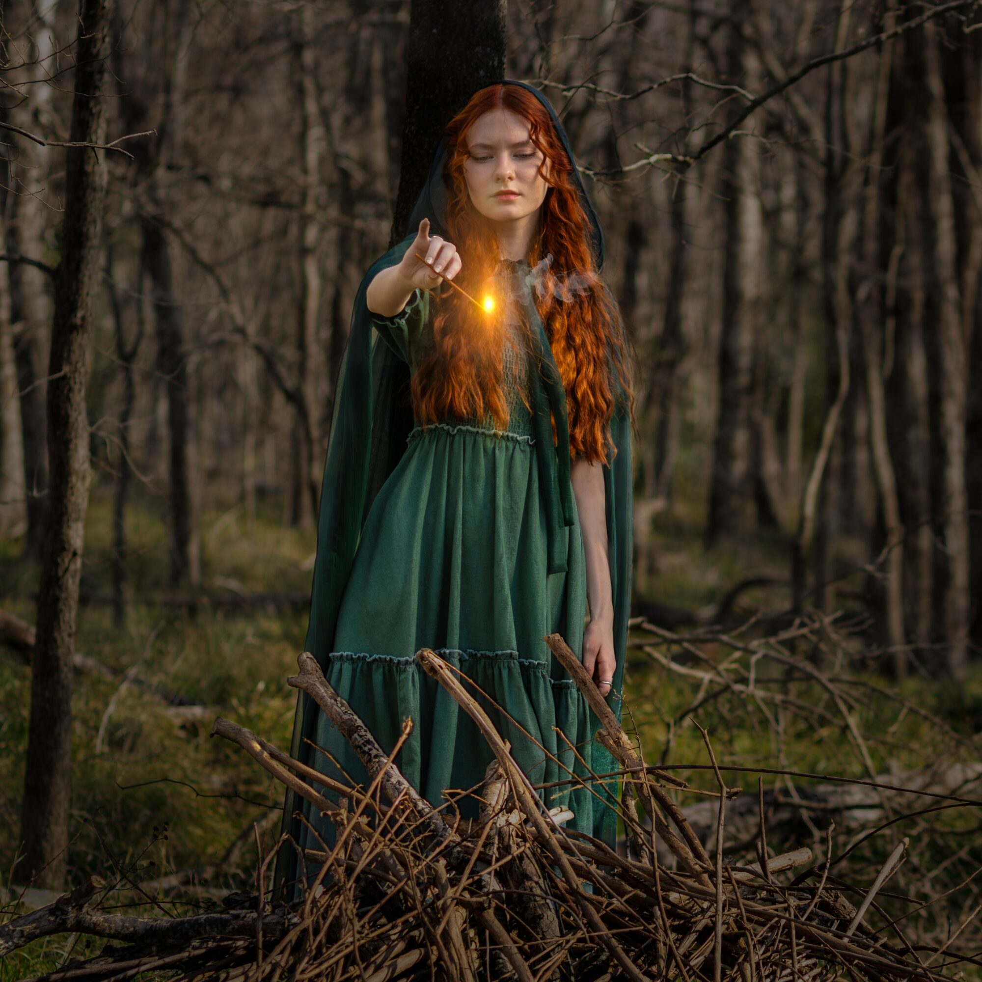Woman with long red hair in a green dress in a forest, holding a glowing object in her hand.