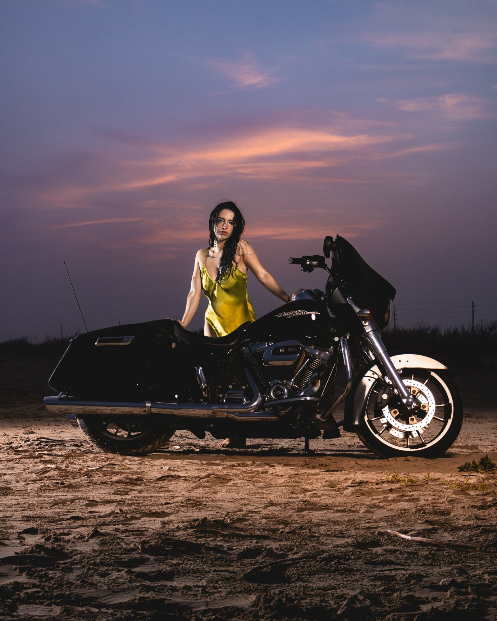 Woman in yellow dress standing next to a black motorcycle outdoors at sunset.