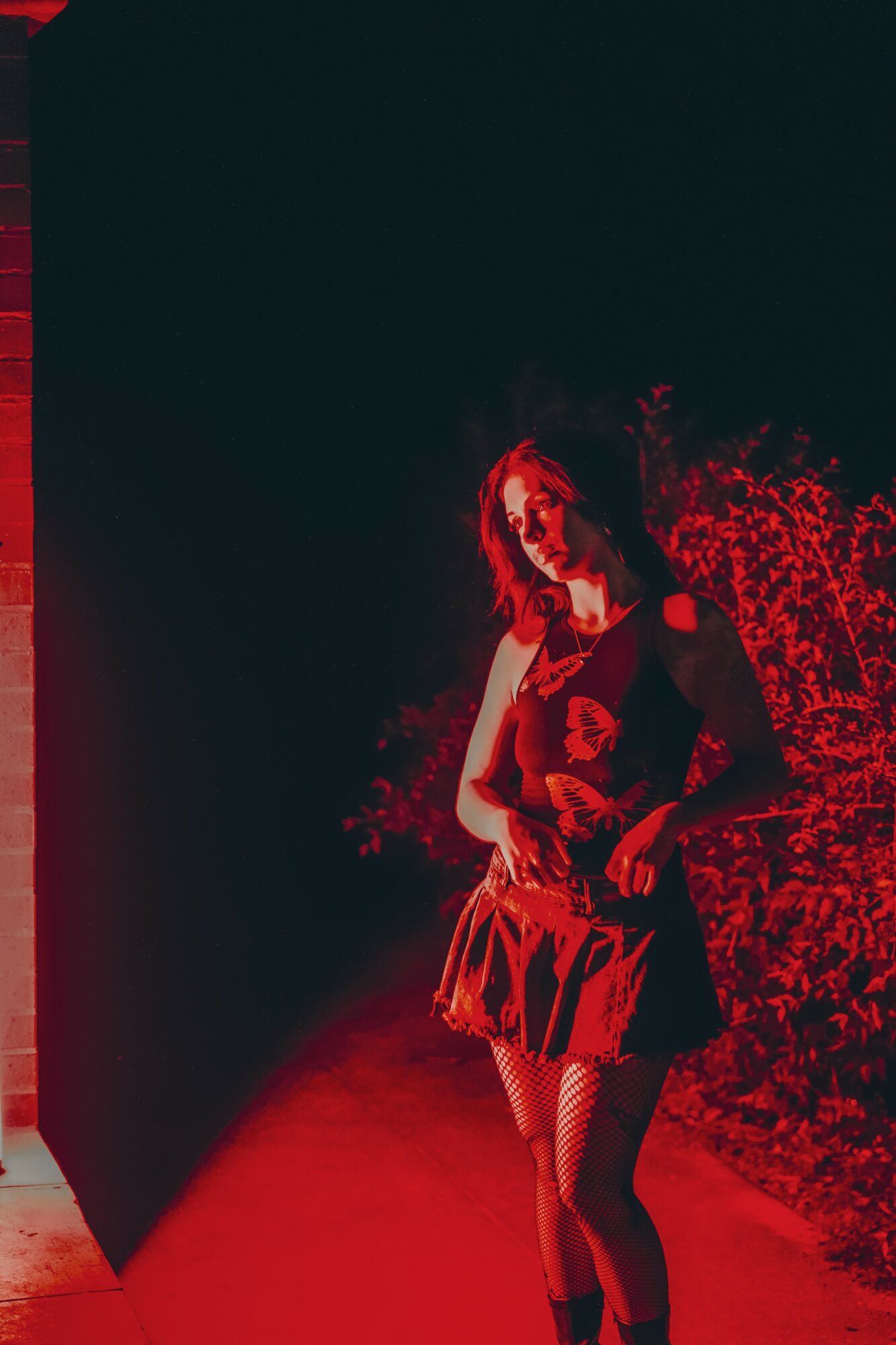 Woman standing outdoors at night, illuminated by red light, with bushes in the background.
