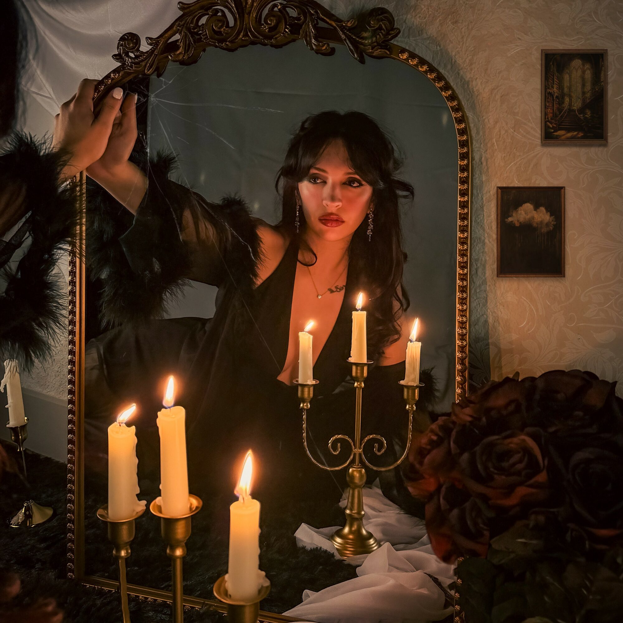 Woman looking into mirror with lit candles and dark flowers nearby, in a dimly lit room with framed pictures on wall.