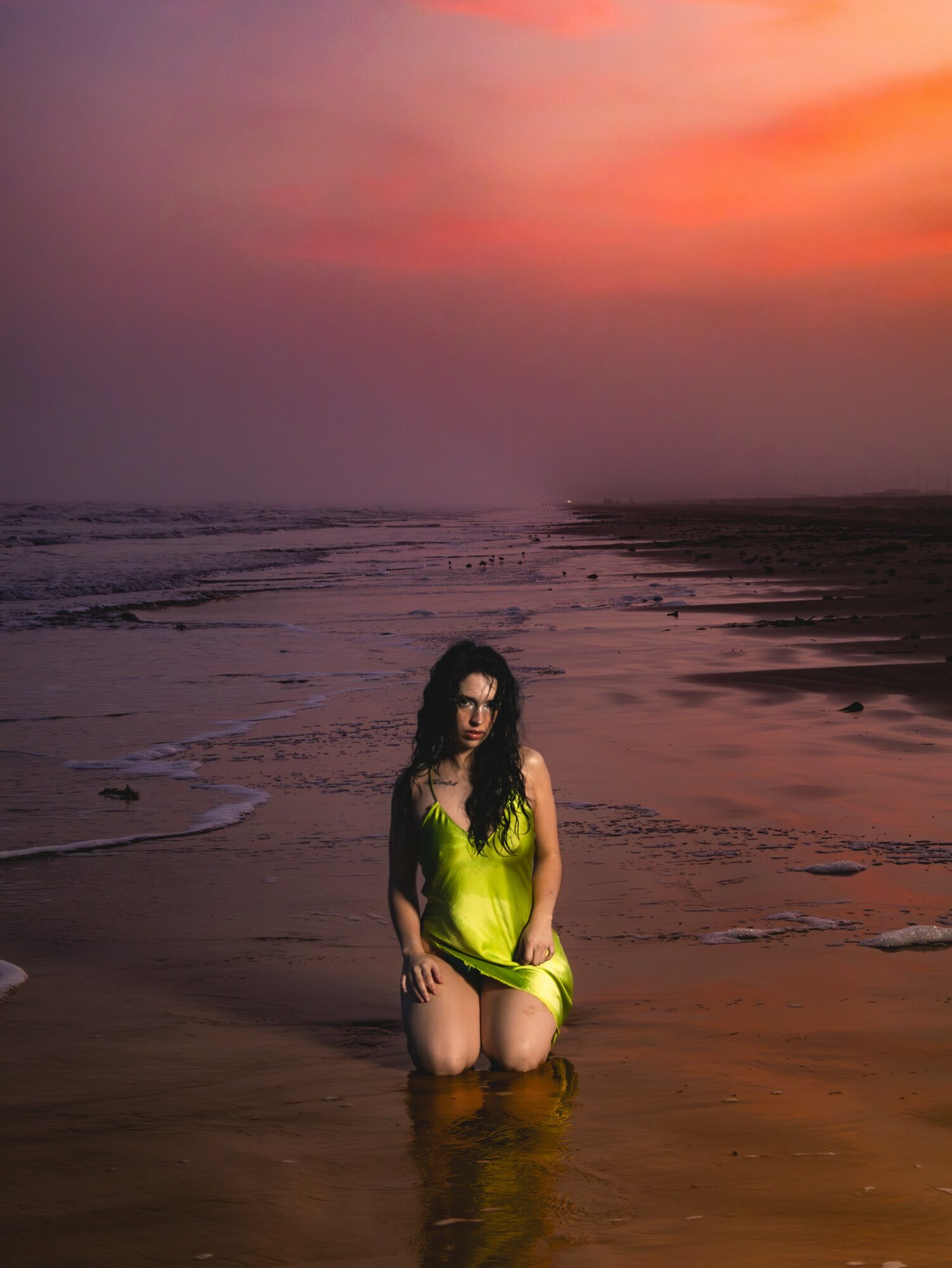 Woman kneeling in shallow water on a beach during sunset, with colorful sky and ocean in background.