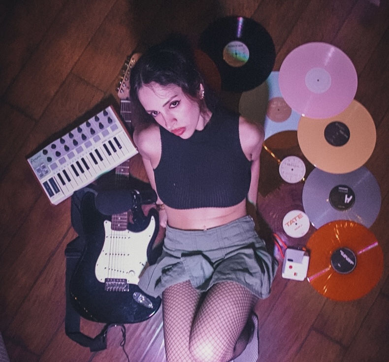 Young woman with dark hair, lying on wooden floor, surrounded by vinyl records, a keyboard, and an electric guitar.