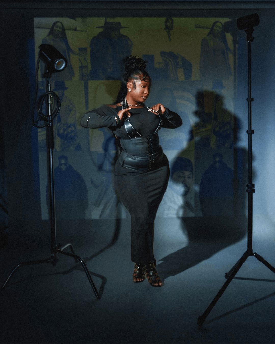 A woman in a black outfit stands in a photography studio with studio lights and a backdrop, adjusting her sleeves.