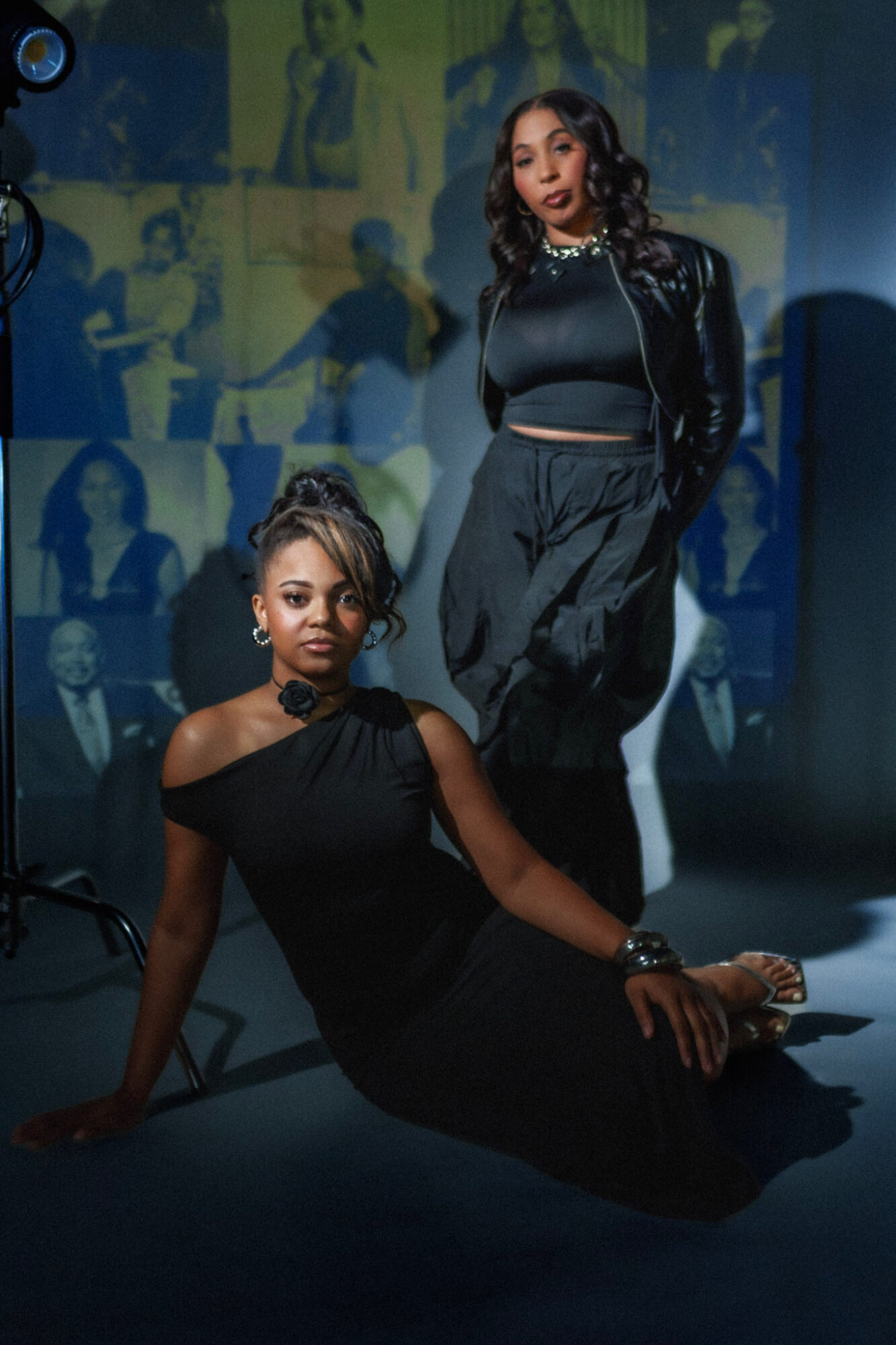 Two women pose in front of a collage of photos, one sitting on the floor and the other standing, both dressed stylishly.