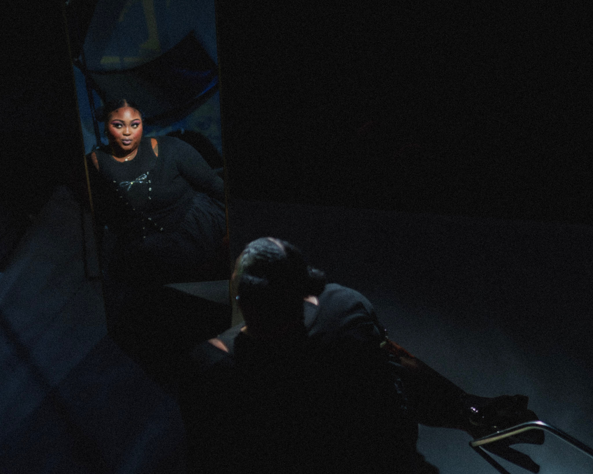 Person looking into a mirror, with a reflection of a woman in dark clothing, in a dimly lit room.