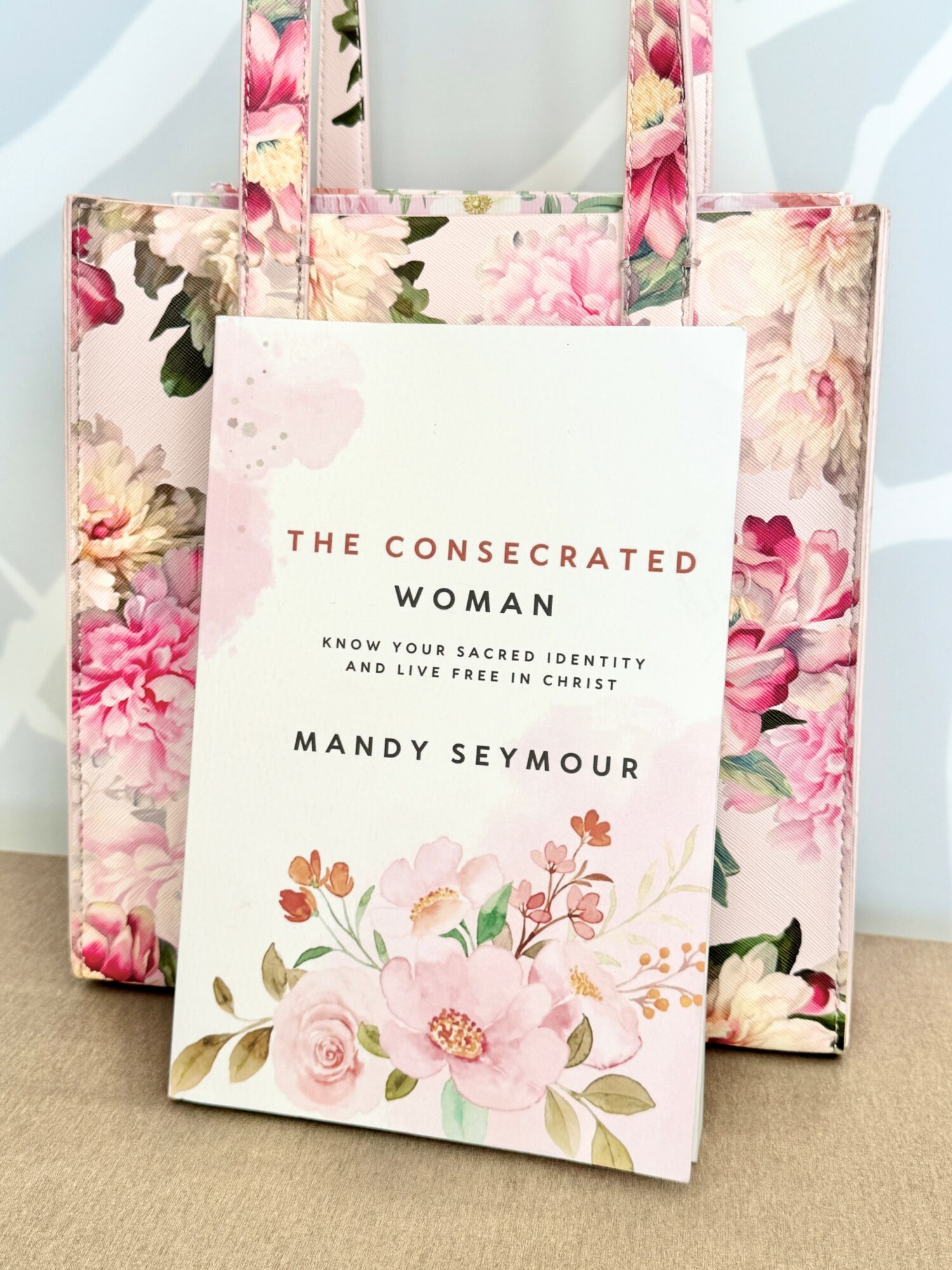 Booklet with floral cover and title about woman and sacred identity, placed in front of a floral bag.