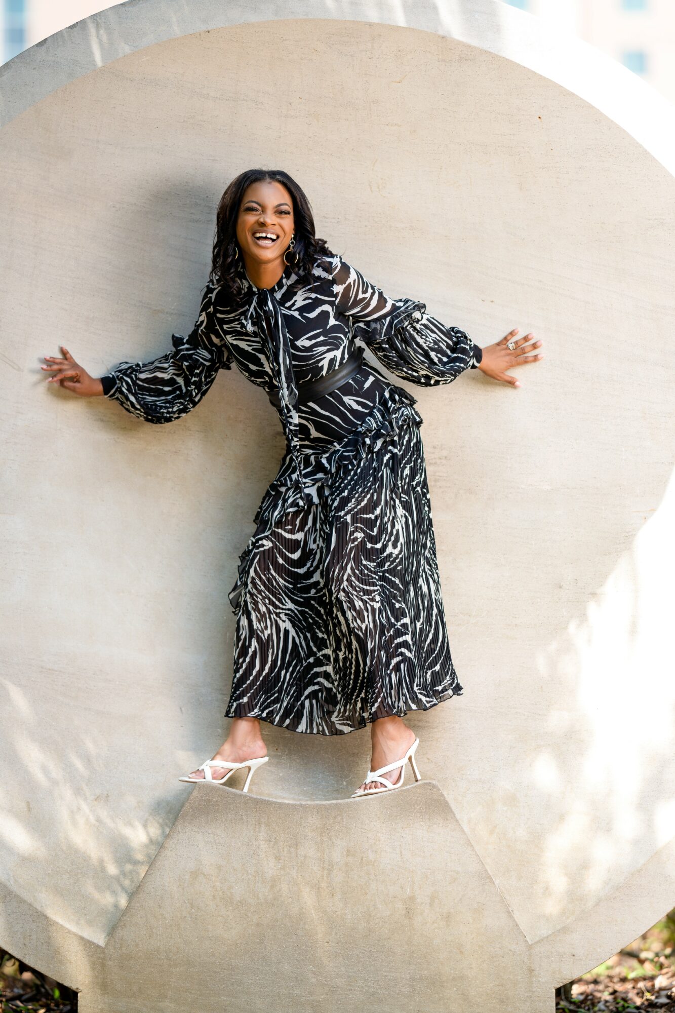 Woman standing with arms outstretched, smiling, wearing a patterned dress, against a circular concrete background.