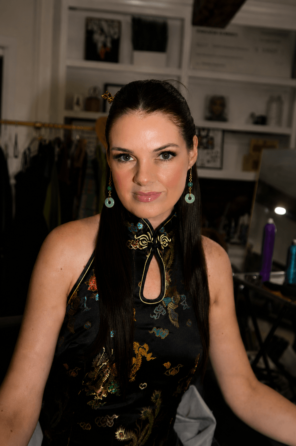 Woman with dark hair and earrings in a room with shelves and clothing in the background.