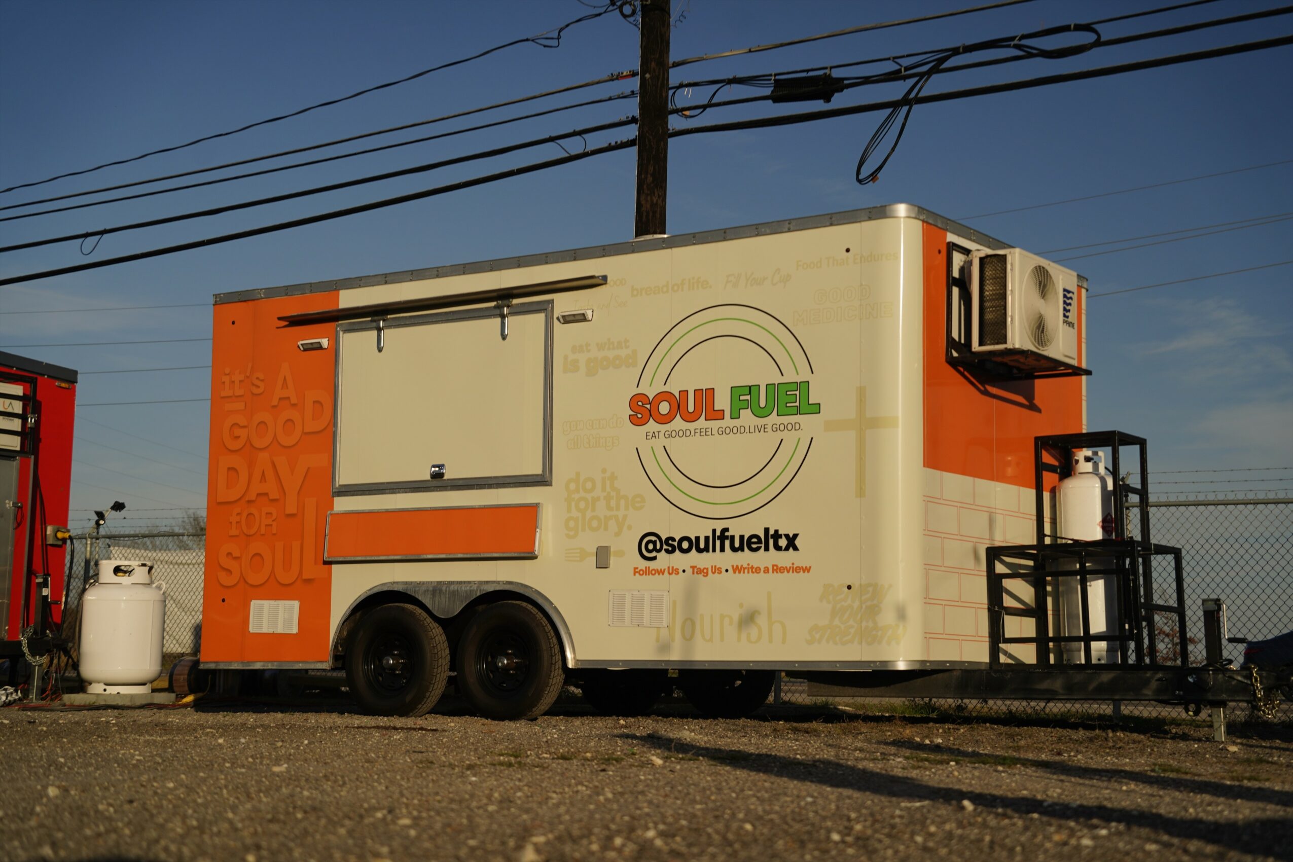 Mobile food truck with orange and white design, logo, and social media handle, parked outdoors with power lines overhead.