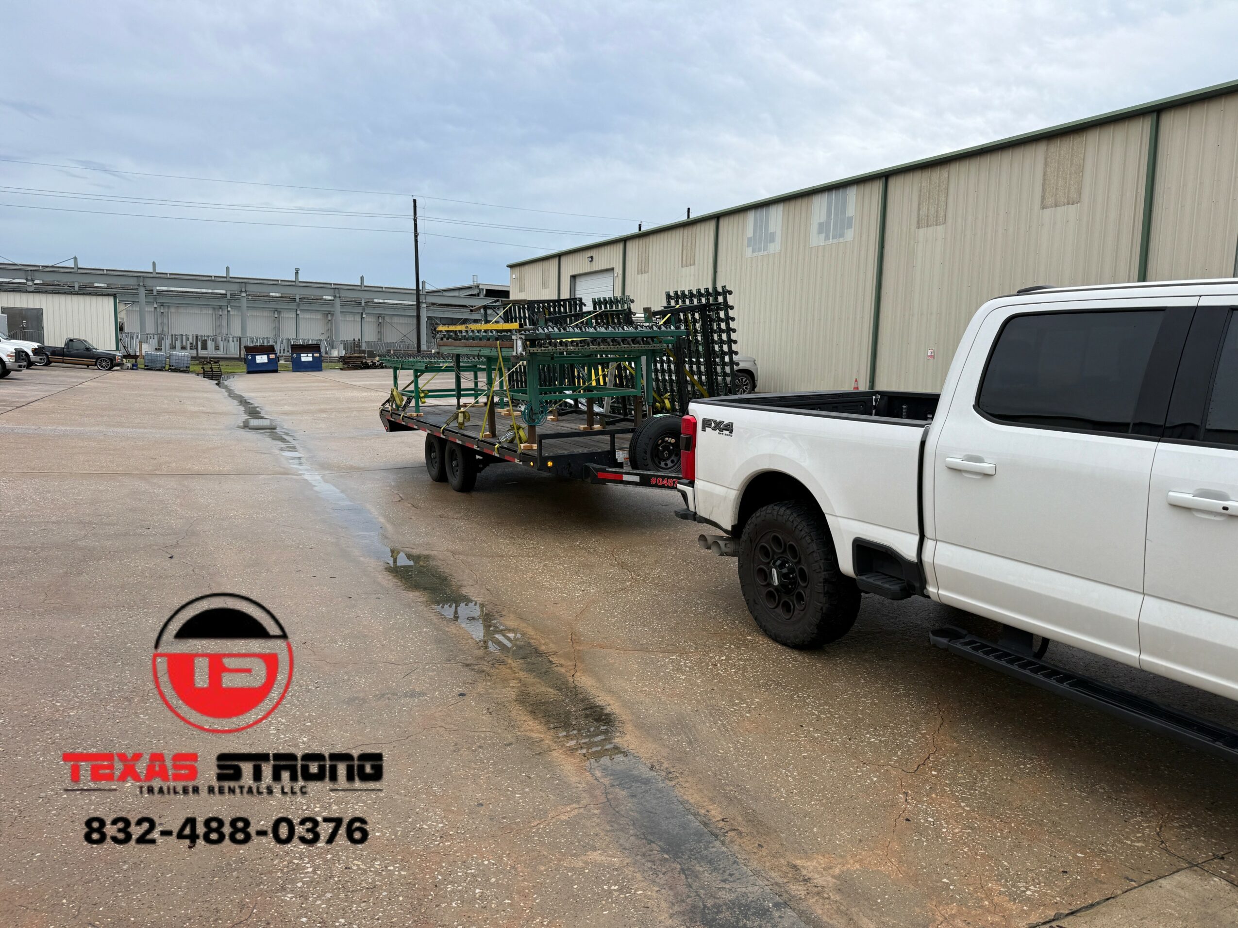 White pickup truck parked next to a trailer with green equipment, in front of a large industrial building, cloudy sky.