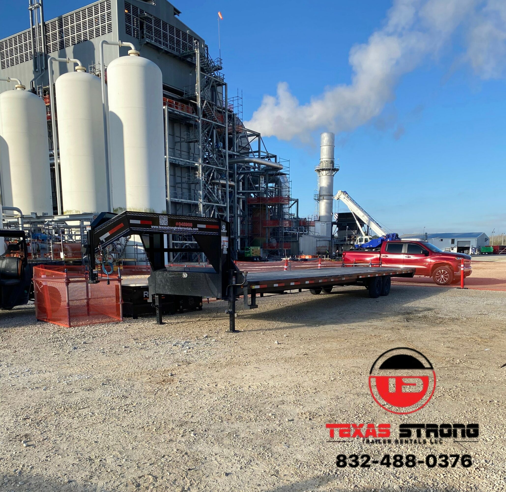 Industrial building with tanks, a red truck, and a flatbed trailer on gravel, with a blue sky and clouds.