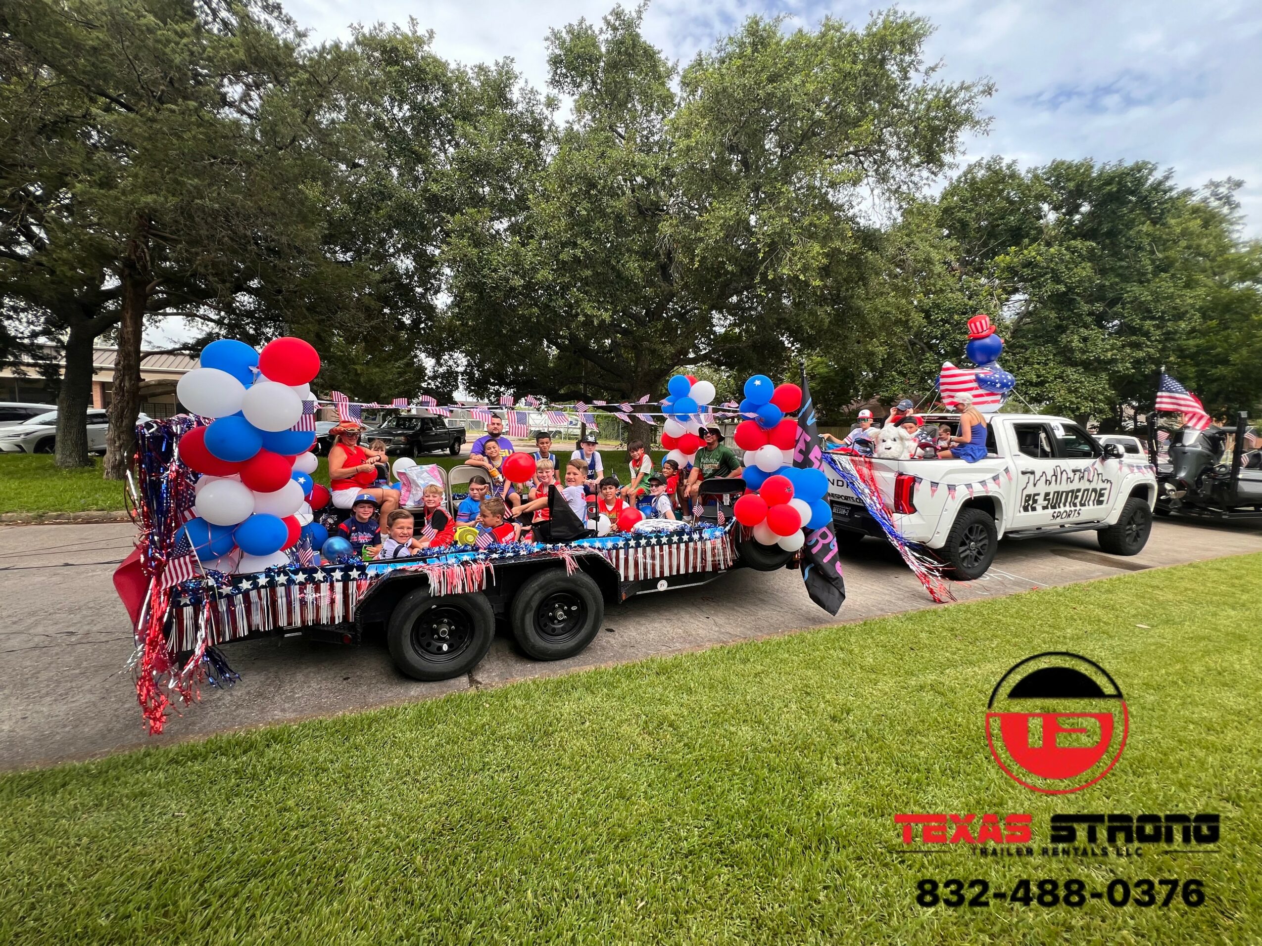 A decorated trailer with balloons and patriotic decorations, attached to a white police vehicle, parked on a street with trees and grass.
