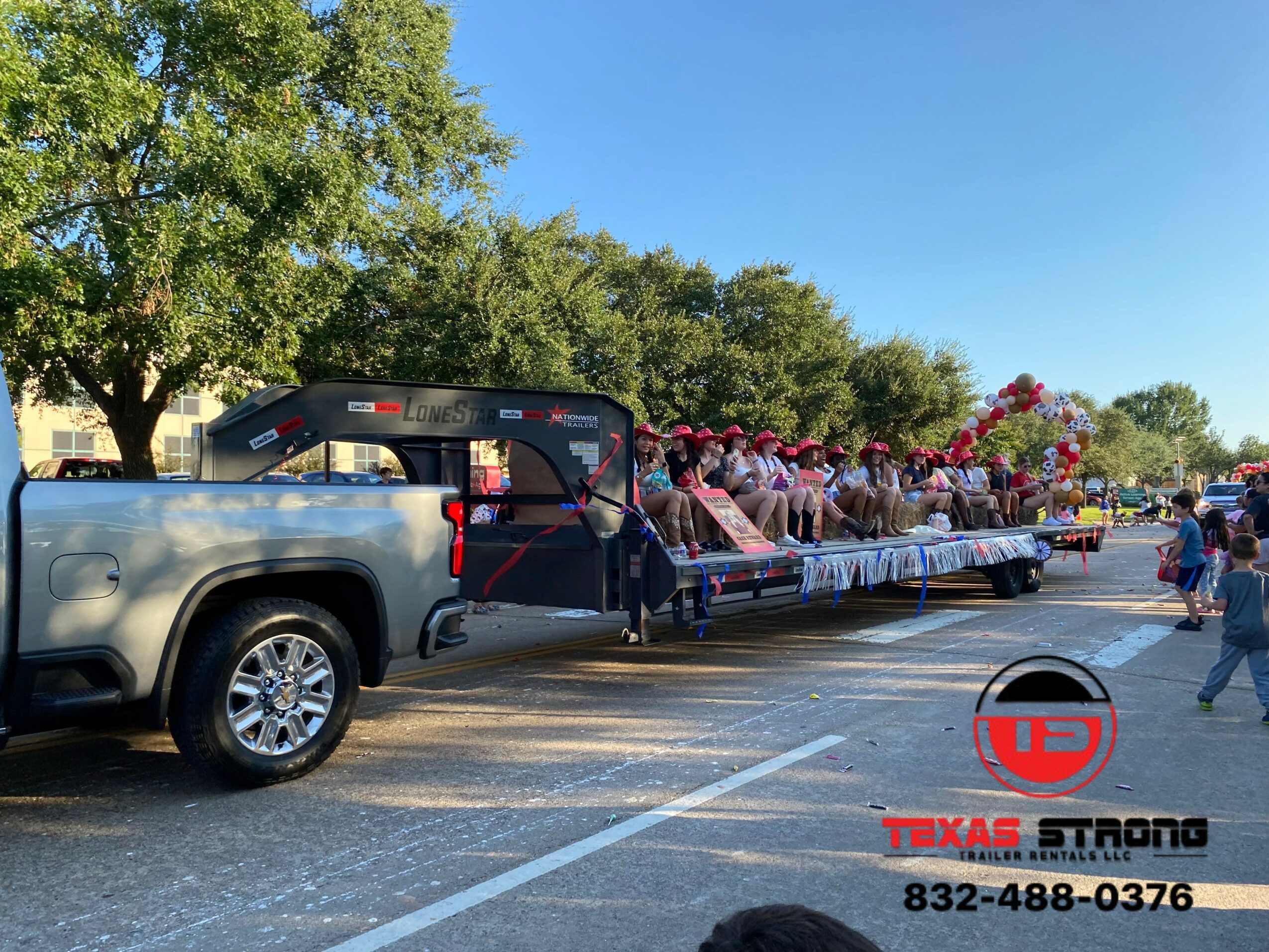 A parade float with people in pink hats and shirts, pulled by a gray pickup truck on a sunny street with trees.