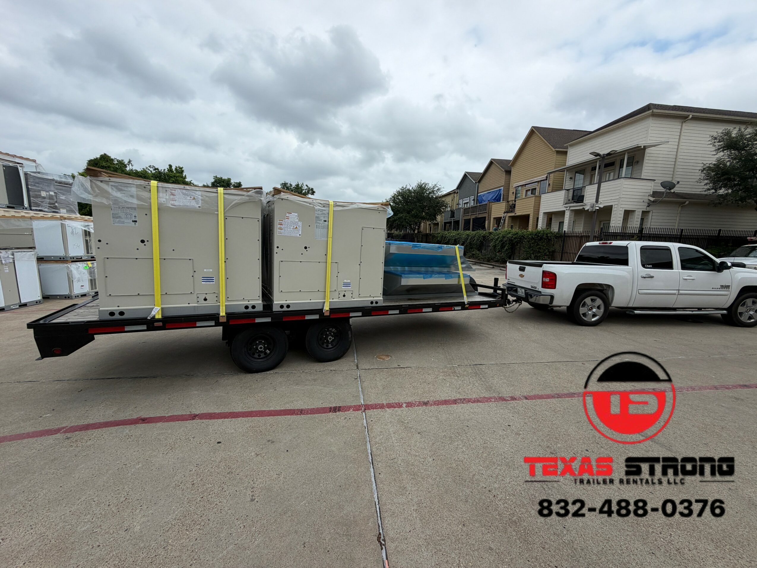 Flatbed trailer with large white equipment, parked in a lot with a white pickup truck nearby, under cloudy sky.