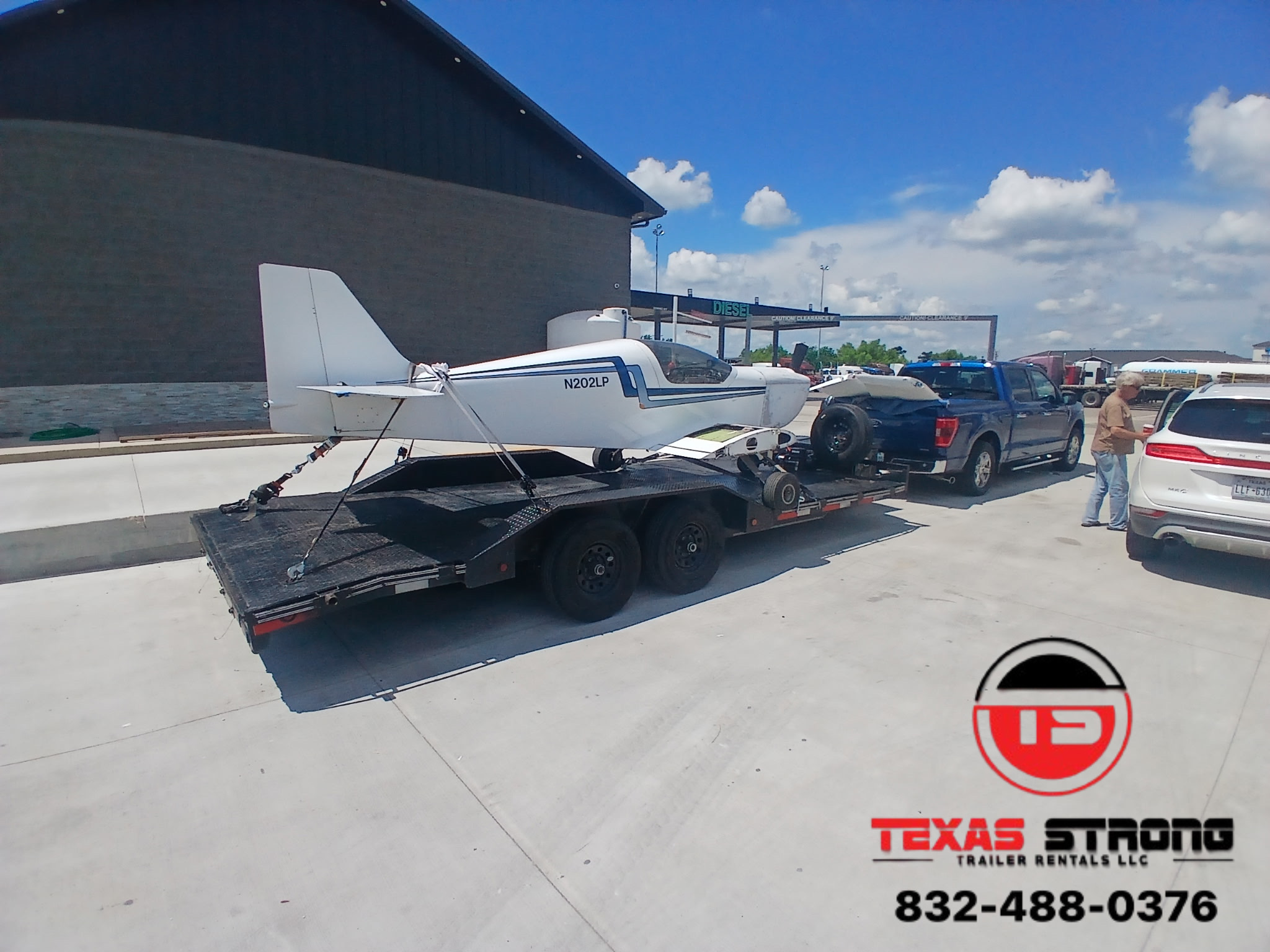 Small white aircraft on a trailer in a parking lot with cars and a building in the background.