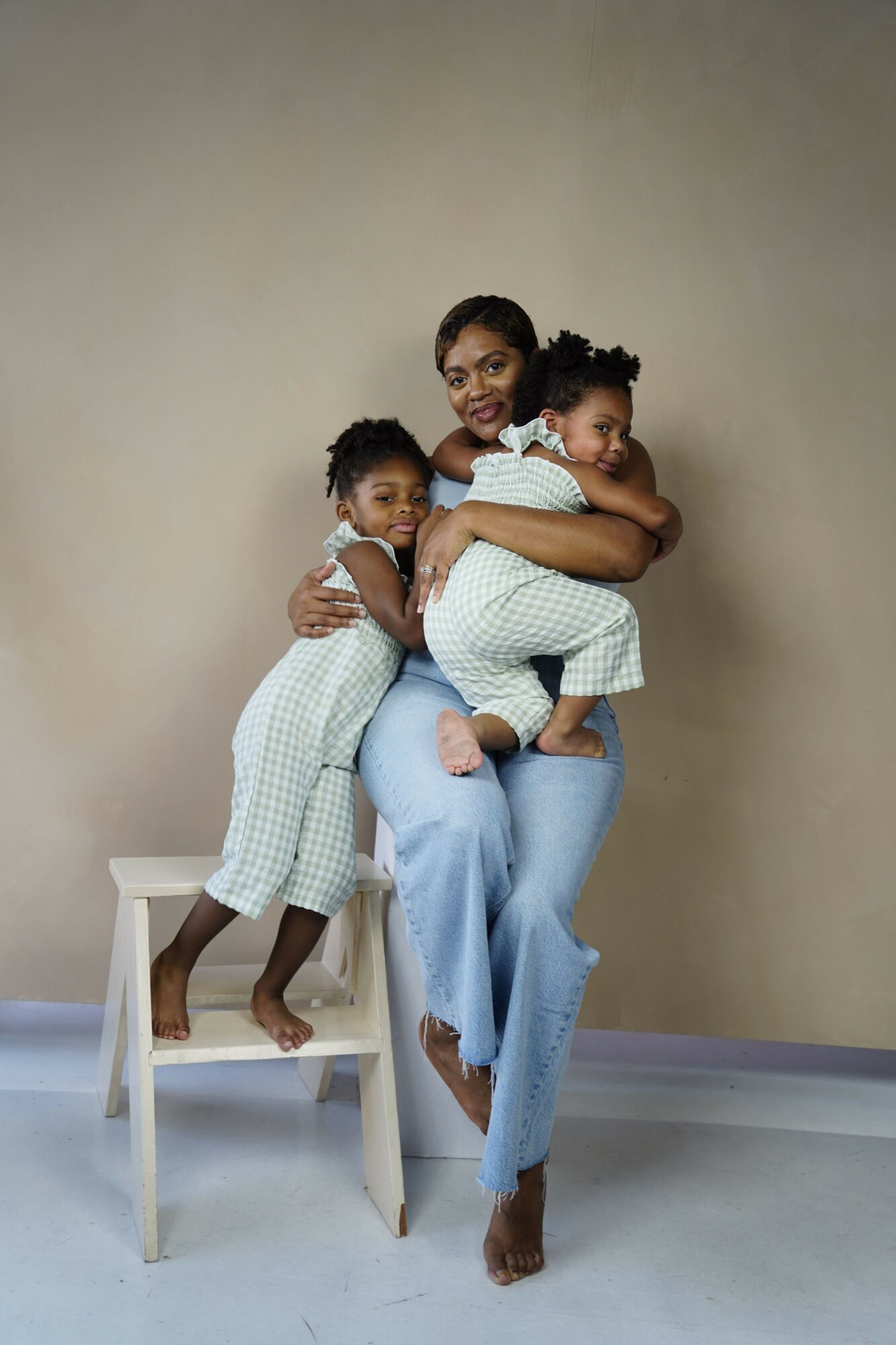 A woman holds a child while another child stands beside them, all smiling and looking at the camera, against a plain background.