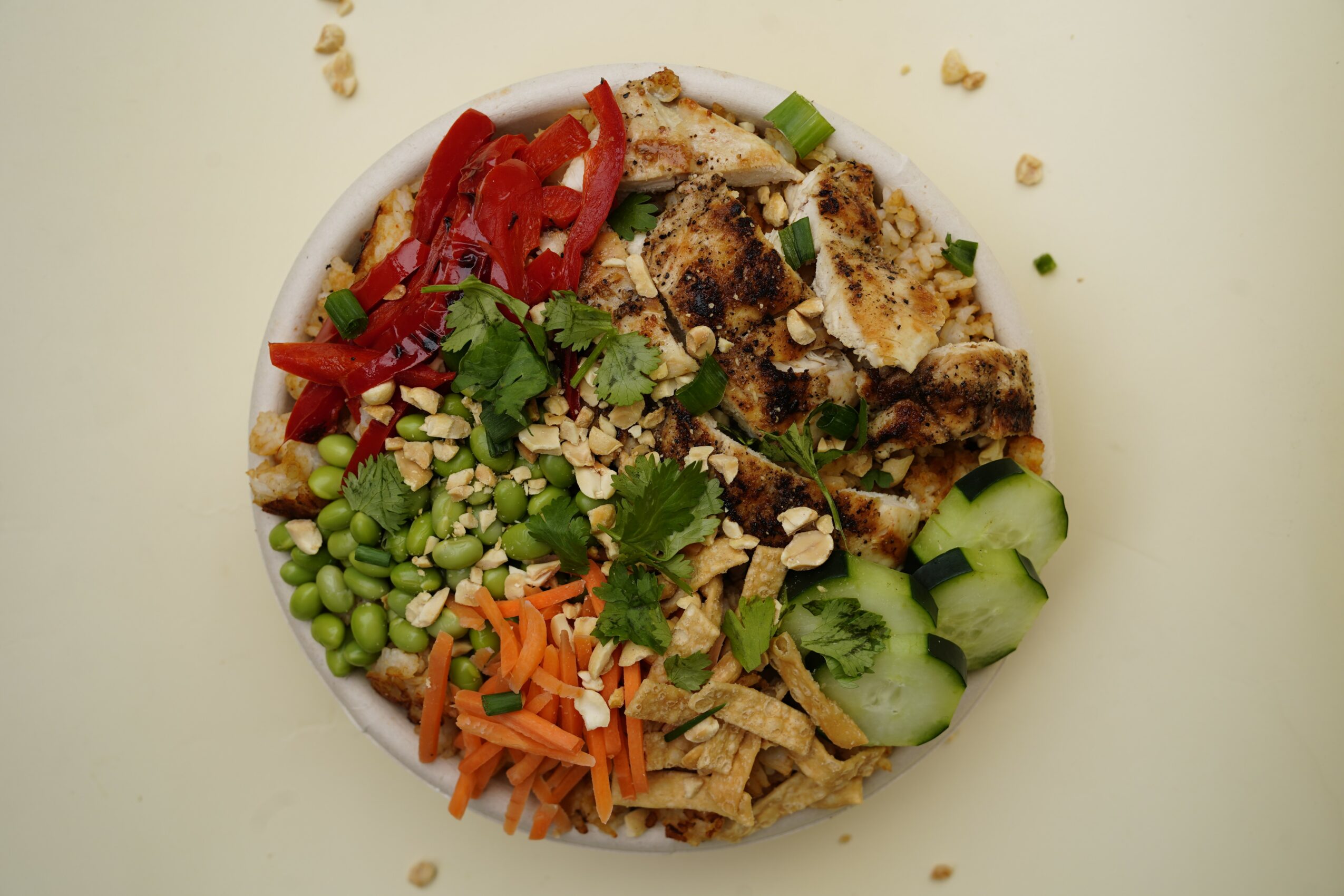 Bowl with grilled chicken, sliced cucumbers, shredded carrots, chopped green onions, red peppers, peas, and cilantro, on a white background.