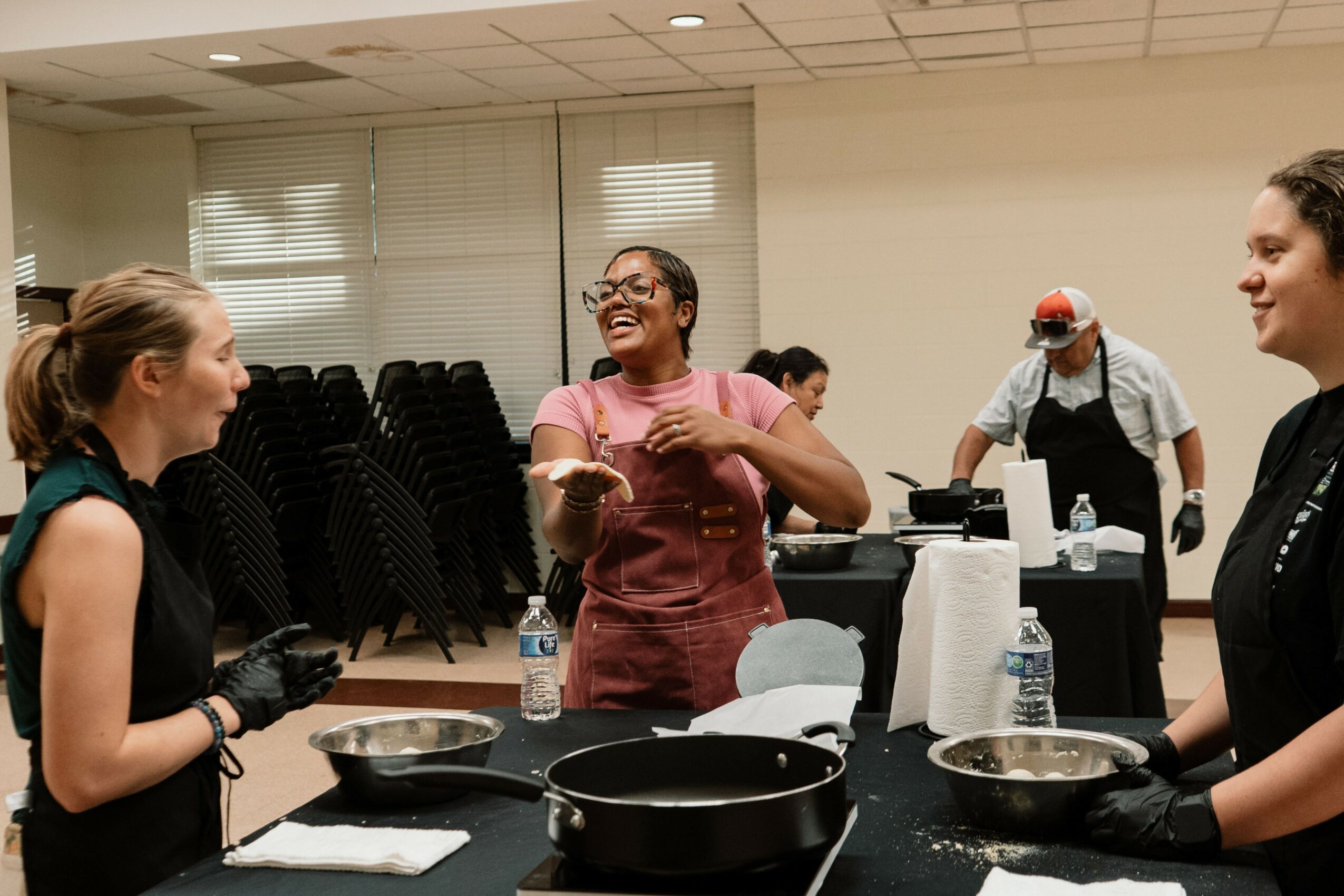 Three women in a kitchen or cooking class, smiling and talking, with cooking utensils and ingredients on the table.