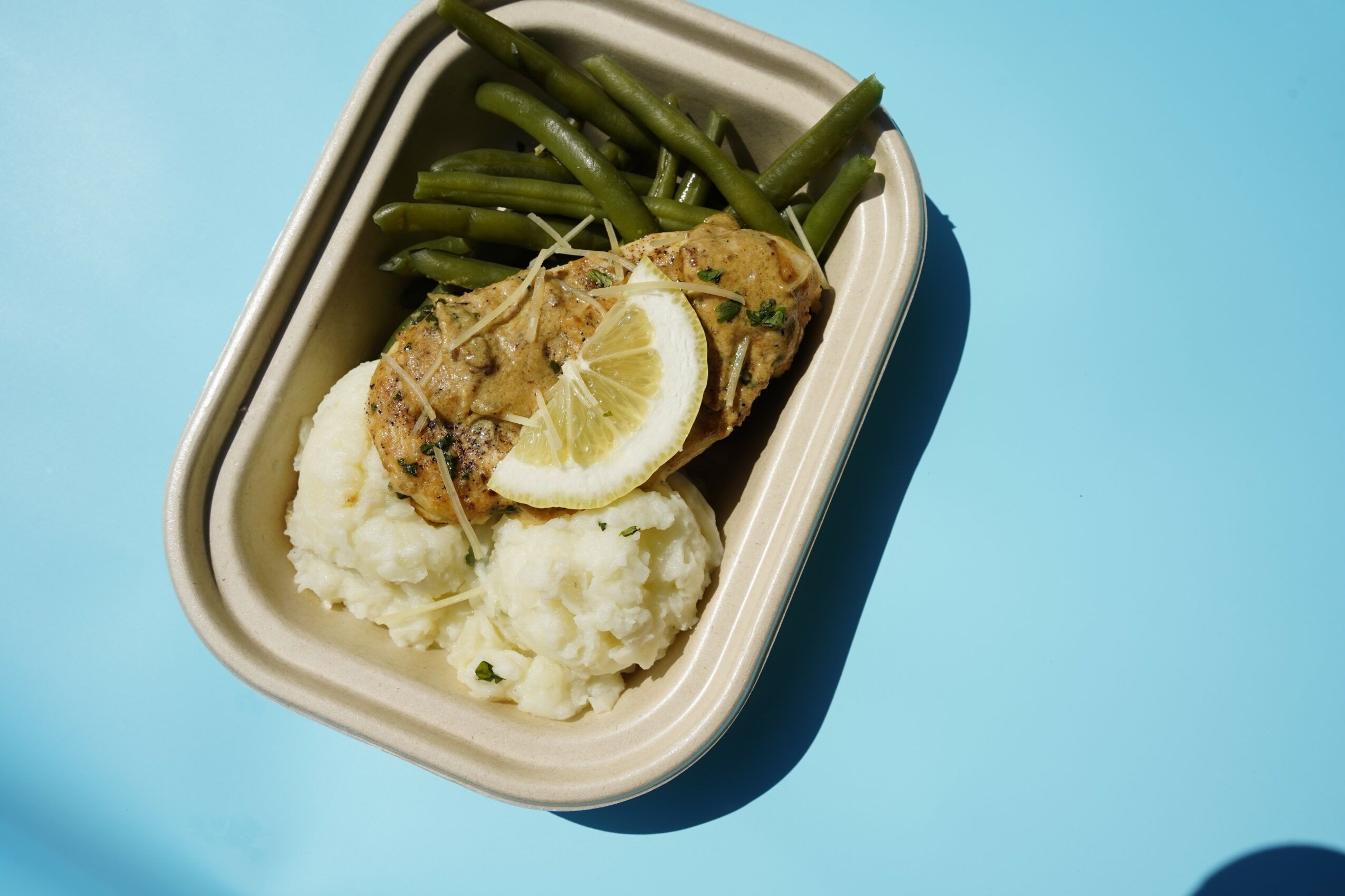A dish with fish, lemon, green beans, and mashed potatoes in a white container on a blue background.