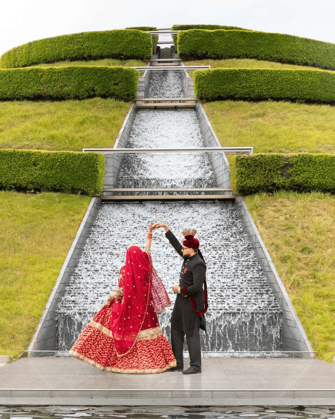 Two people in traditional attire stand near a cascading water feature in a landscaped garden with trimmed hedges and steps.