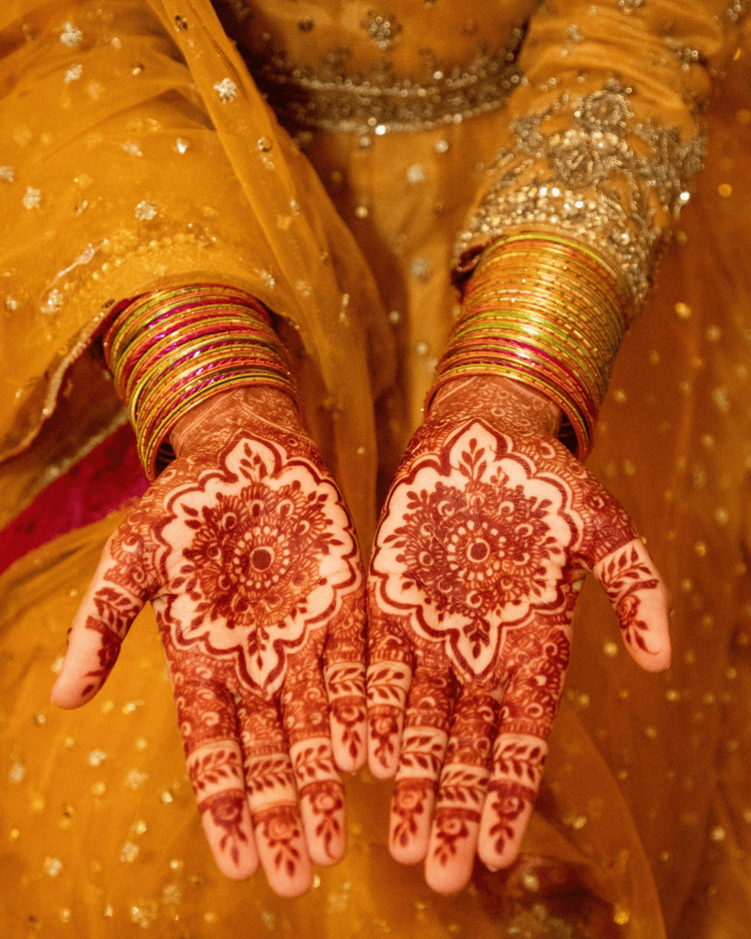 Person's hands with intricate henna designs, wearing multiple gold bangles, in yellow traditional attire with embellishments.