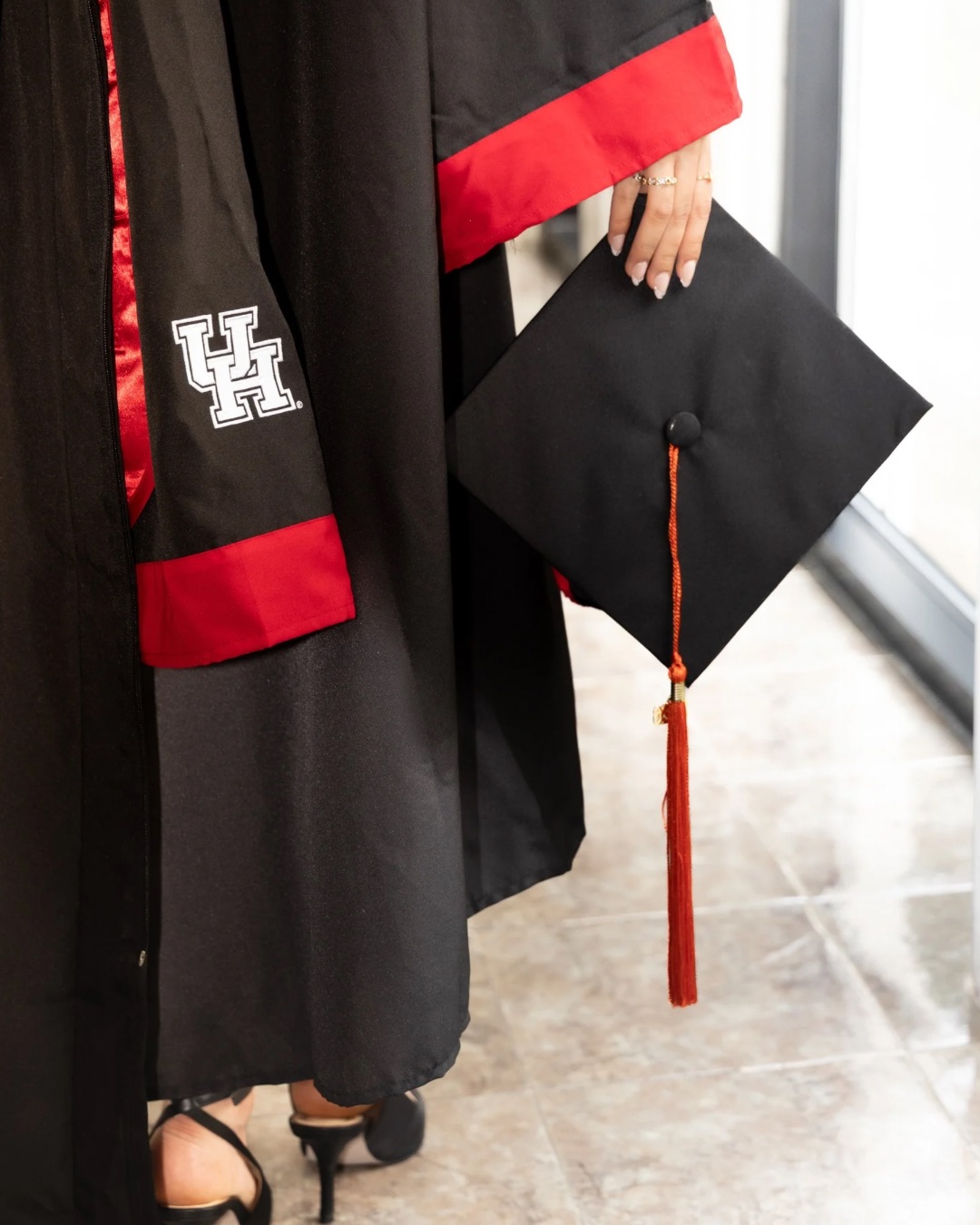 Person in graduation gown holding a cap with a tassel, standing indoors near a window.