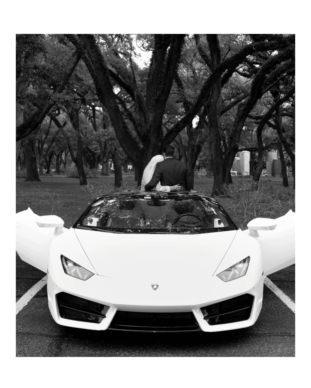 Person leaning over a white sports car parked outdoors with trees in the background.