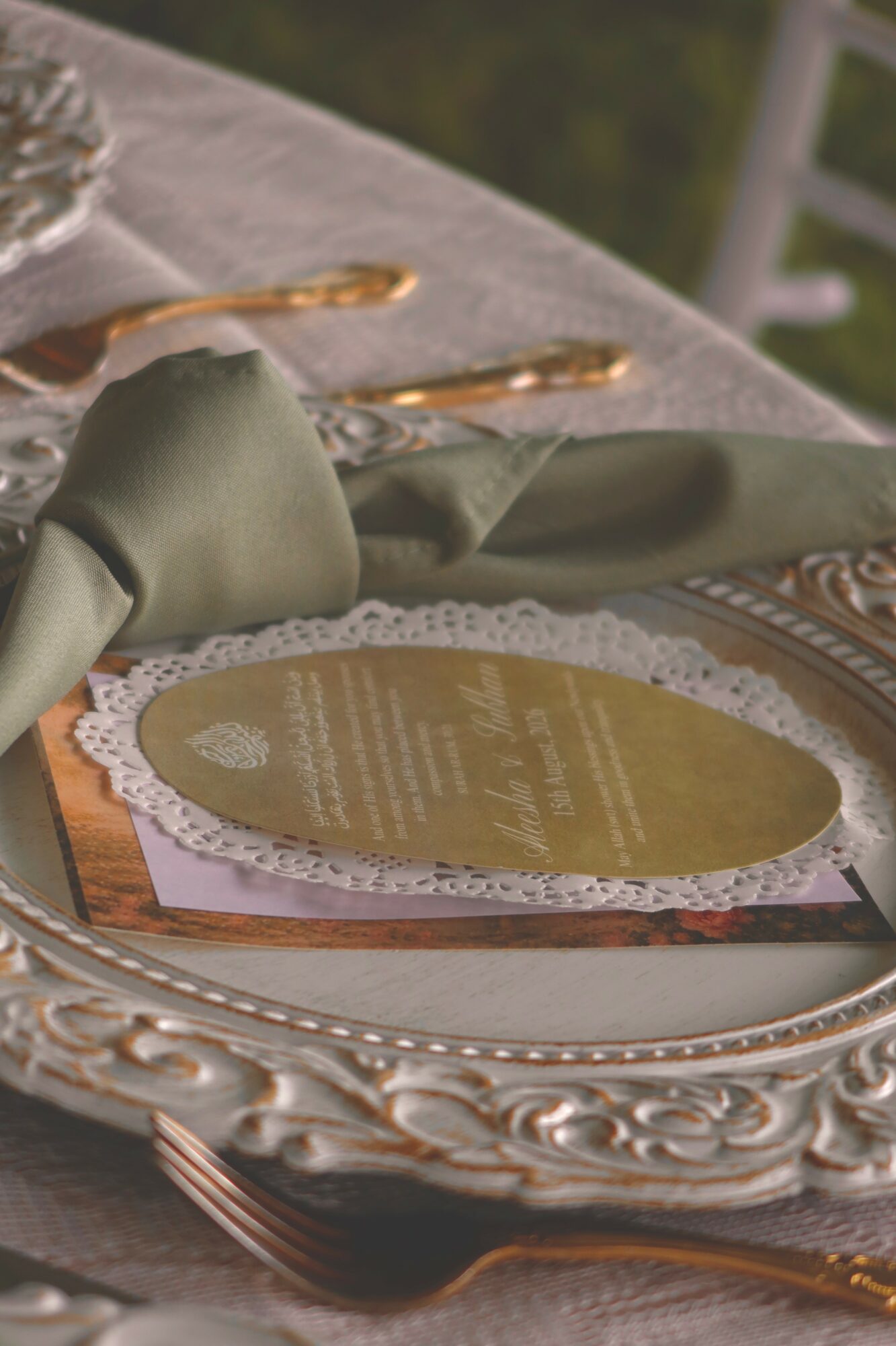 Elegant table setting with a gold-colored menu on a decorative plate, surrounded by gold utensils and a green napkin, outdoors.