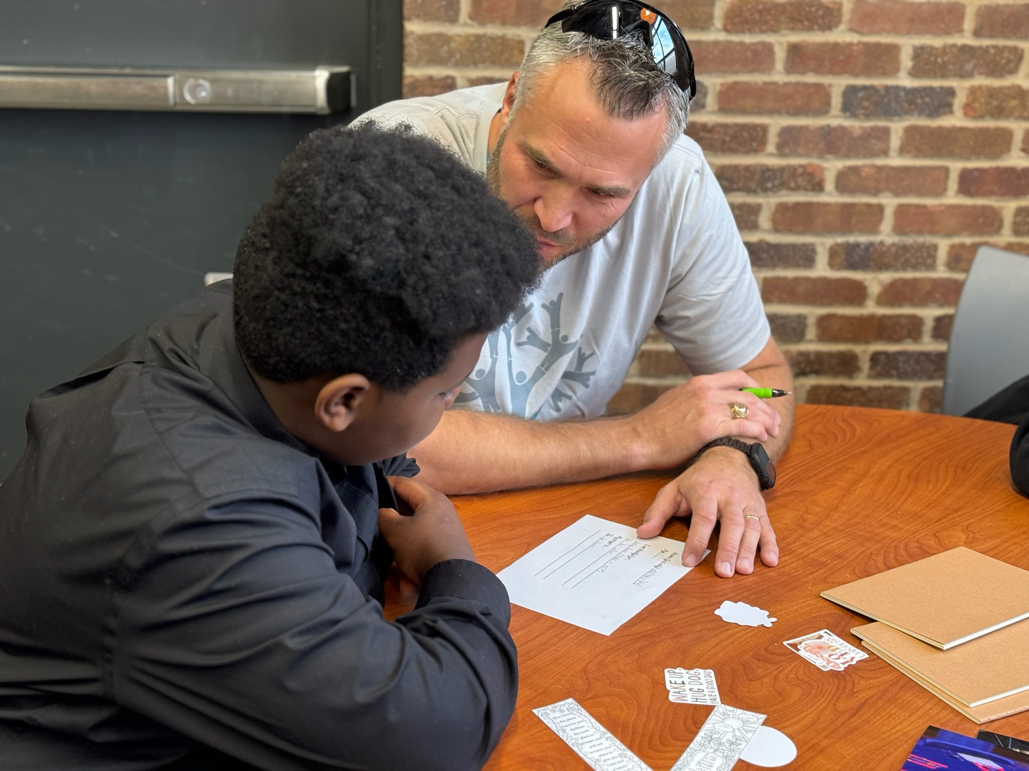 Two men sit at a table with papers and envelopes, engaged in a discussion, in front of a brick wall.