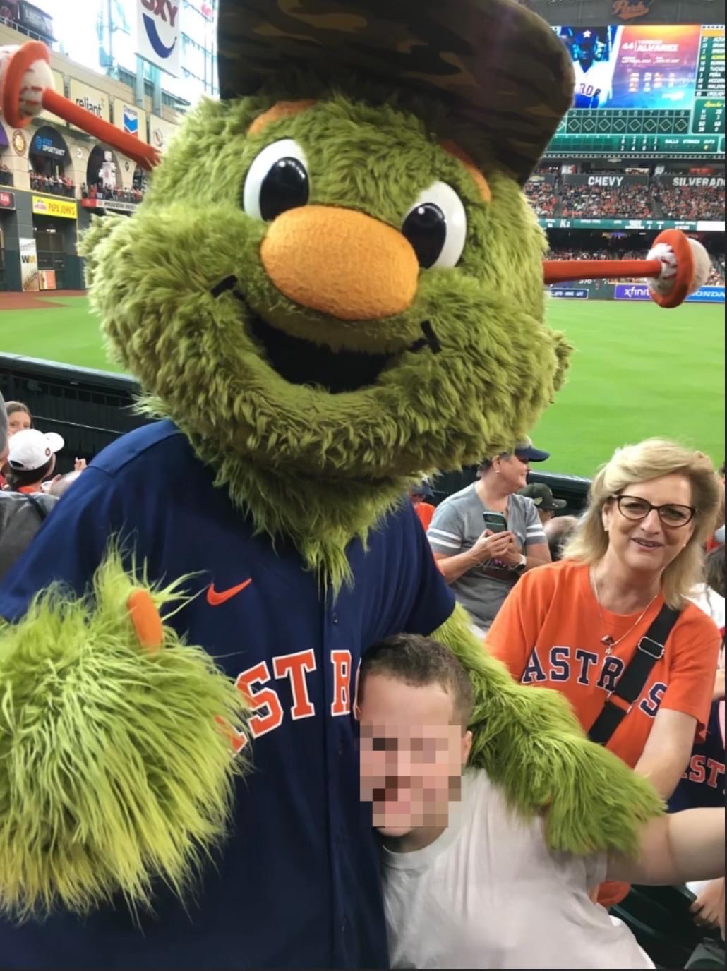 Mascot with green fur, wearing a baseball cap and a blue jersey, standing next to a smiling woman in an orange Astros shirt, at a stadium.