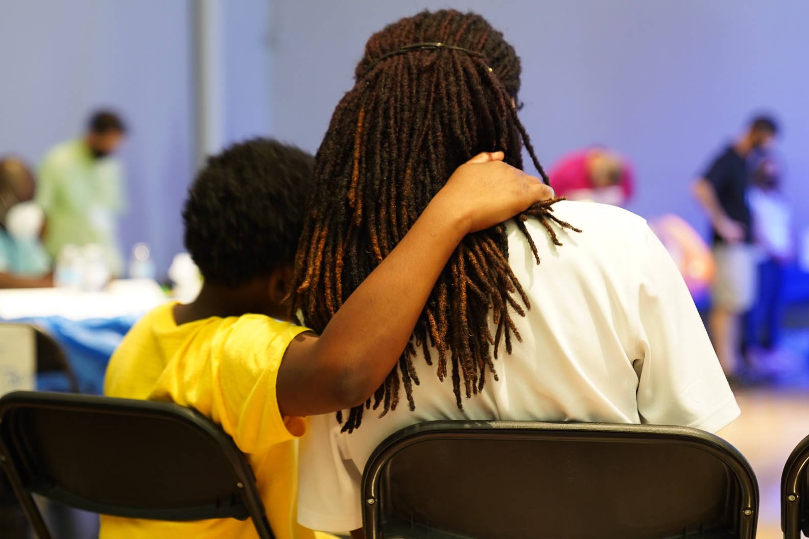 Child with arm around adult, both sitting on chairs, in a room with people and tables in the background.