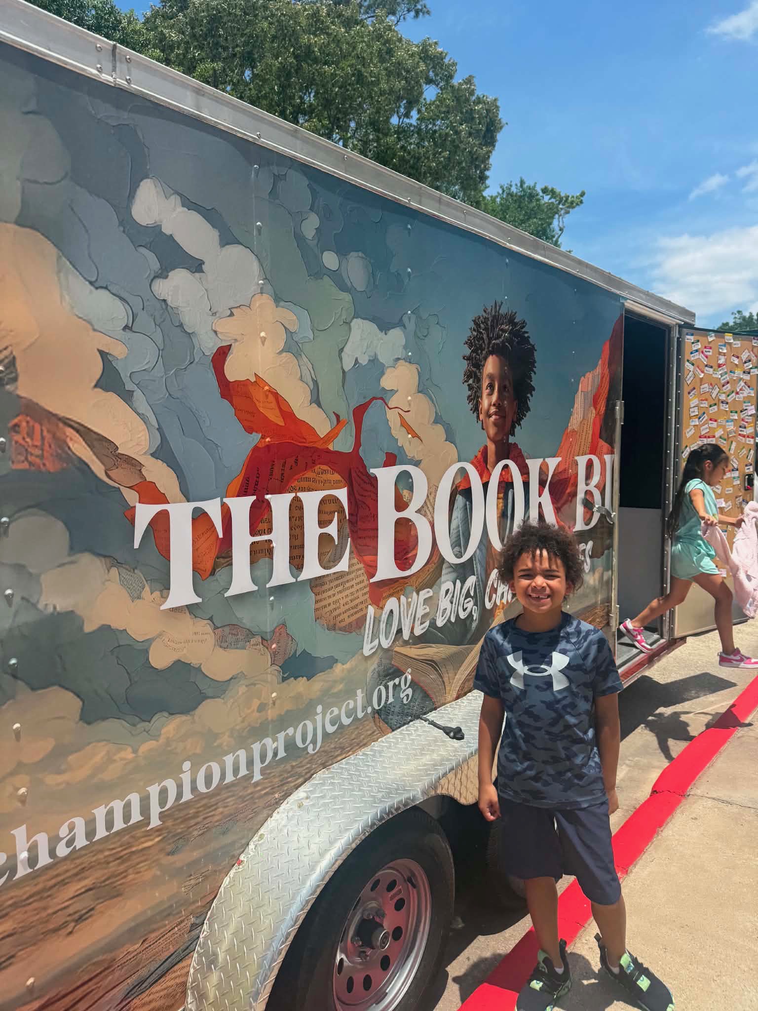 Two children standing in front of a colorful truck with a mural and text, outdoors on a sunny day.