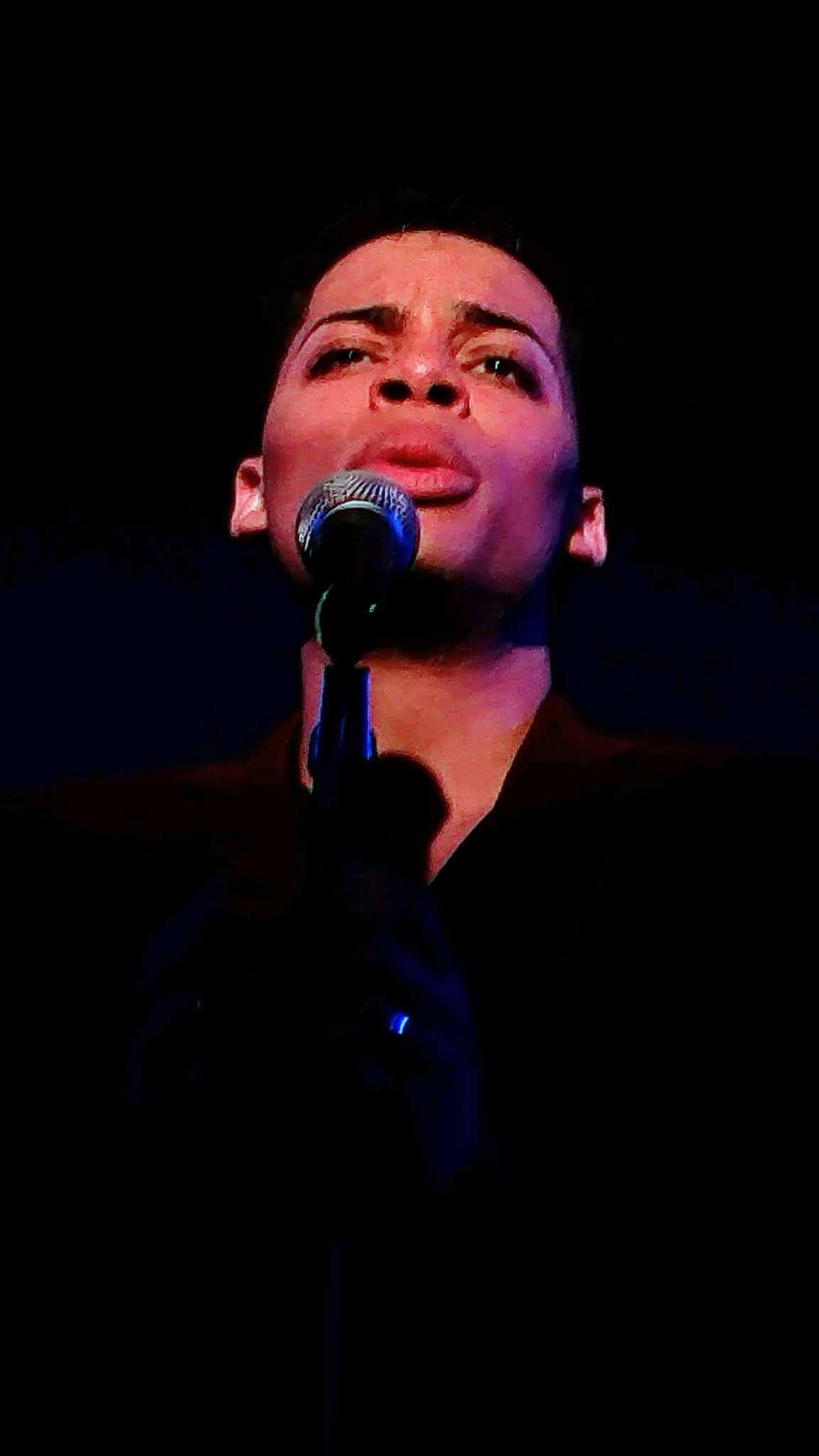 Person singing into a microphone with a serious expression, against a dark background.