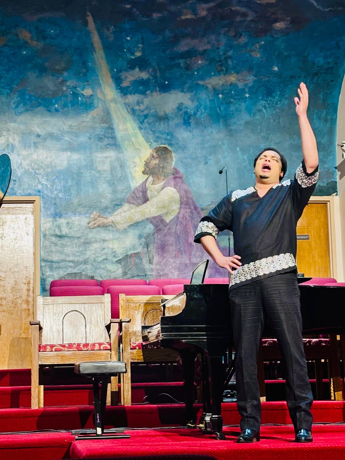 Performer singing with one arm raised on stage, colorful background with a painted scene, piano nearby, red carpeted stage.