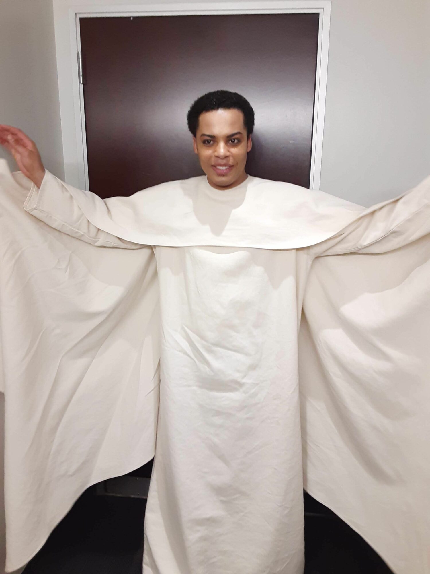 Person standing in a hospital bed with arms outstretched, wearing a white gown, smiling, with a monitor behind.