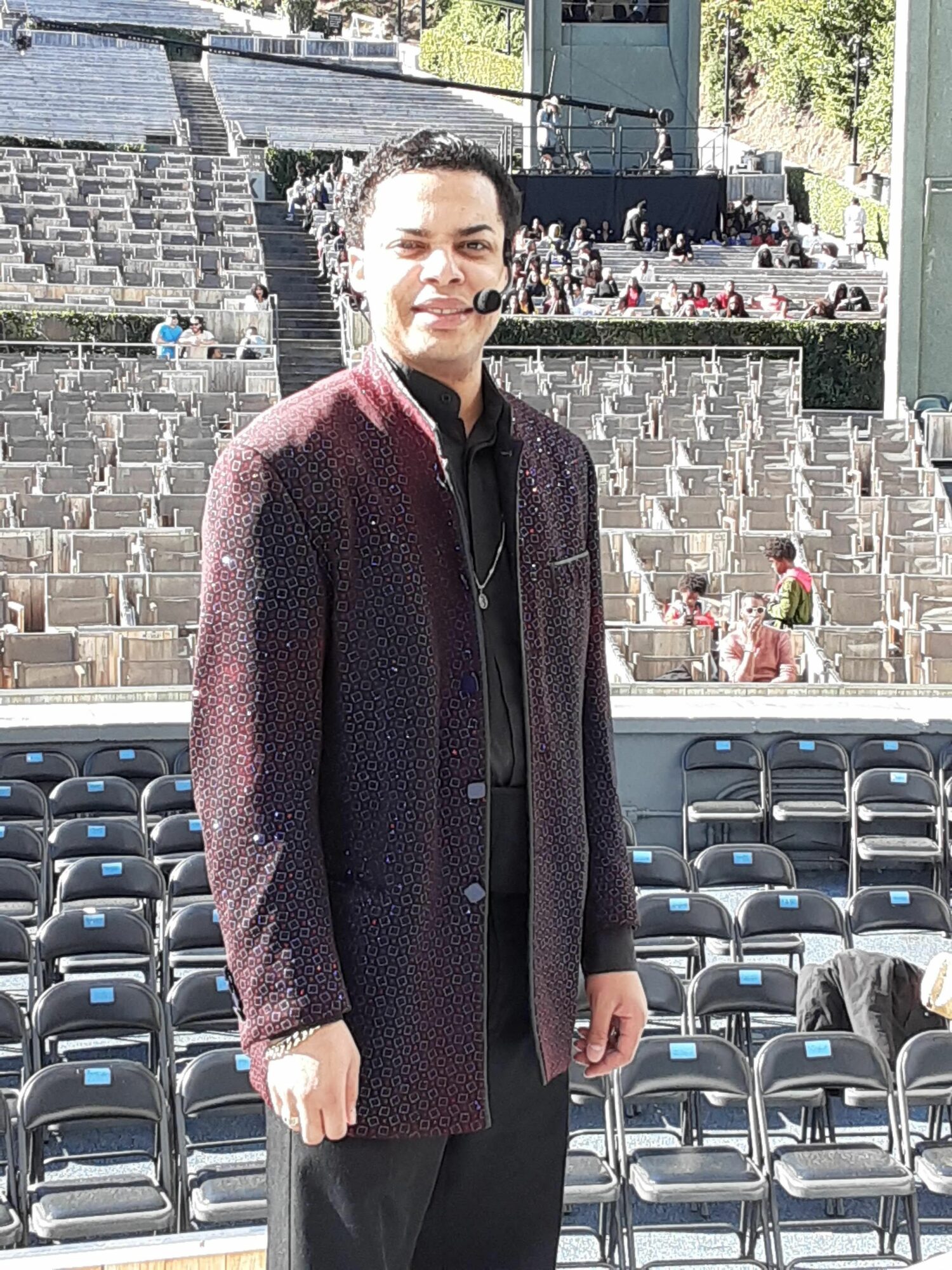 Woman standing outdoors near empty chairs and a brick wall, wearing a dark coat and pants.